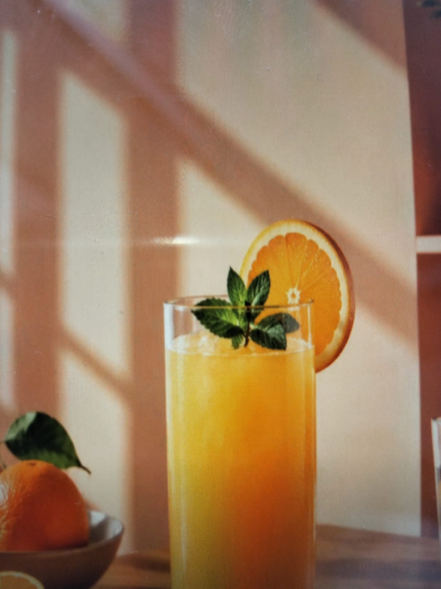 A tall glass filled with a vibrant orange beverage, possibly orange juice or a cocktail, is garnished with a fresh mint sprig and a slice of orange on the rim. The drink appears to be blended or crushed ice based, with a frothy texture. To the left of the glass, a small bowl contains a whole orange and a green leaf, suggesting the beverage is made from fresh fruit. The background is a warm, neutral tone with soft shadows cast by an unseen light source, creating a gentle ambiance. The scene is set on a wooden surface, indicating an indoor setting. The image does not contain any specific information to link it to Payagaboh, Indonesia, or any particular city.