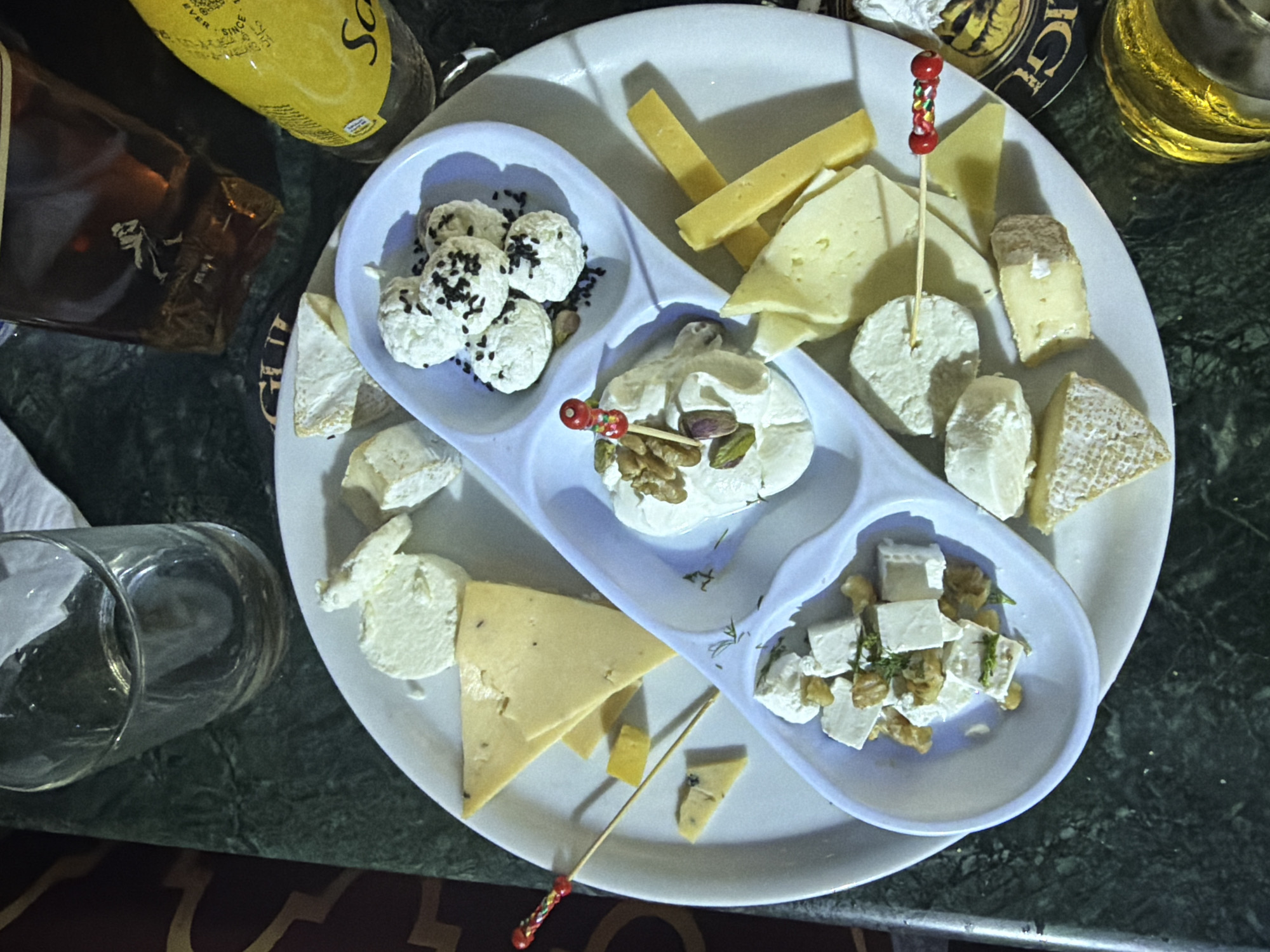 An overhead, close-up shot shows a cheese platter and drinks on a dark green, marble-patterned table, likely in a restaurant in Sharm El Sheikh, Egypt. The platter features various cheeses, including a soft, creamy cheese topped with black seeds, cubed feta cheese mixed with nuts and herbs, and several wedges of hard and soft cheeses, some with visible rind. The cheeses are arranged in a white, multi-compartment dish. A cocktail pick with red and green beads pierces one of the soft cheeses. To the left, a glass is partially filled with liquid. In the background, a dark bottle with a label depicting a walking figure and a yellow bottle with the word "Schweppes" are visible, along with a bottle cap with a gold and black label. The lighting is dim, creating a warm and intimate atmosphere.
