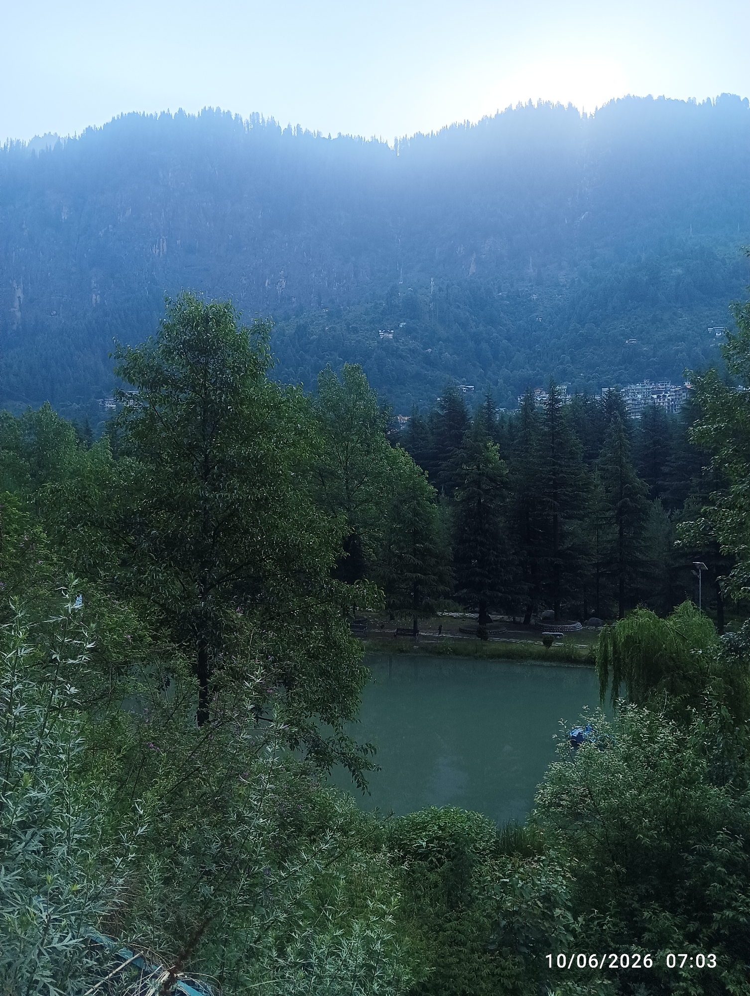 This is a vertical, medium-shot photograph of the scenic Vashisht Village in Manali, India, captured at an angle from a slightly elevated position. The scene is dominated by a tranquil lake, its surface reflecting the muted tones of the overcast sky. Lush green foliage, comprising tall trees and dense bushes, frames the lake, creating a naturalistic and serene environment. In the background, a mist-shrouded mountain range, heavily forested with coniferous trees, rises majestically. The presence of scattered buildings on the mountainside suggests the proximity of human habitation. The overall atmosphere is peaceful and idyllic, characteristic of a mountain landscape. The timestamp "10/06/2026 07:03" is visible in the lower right corner of the image.