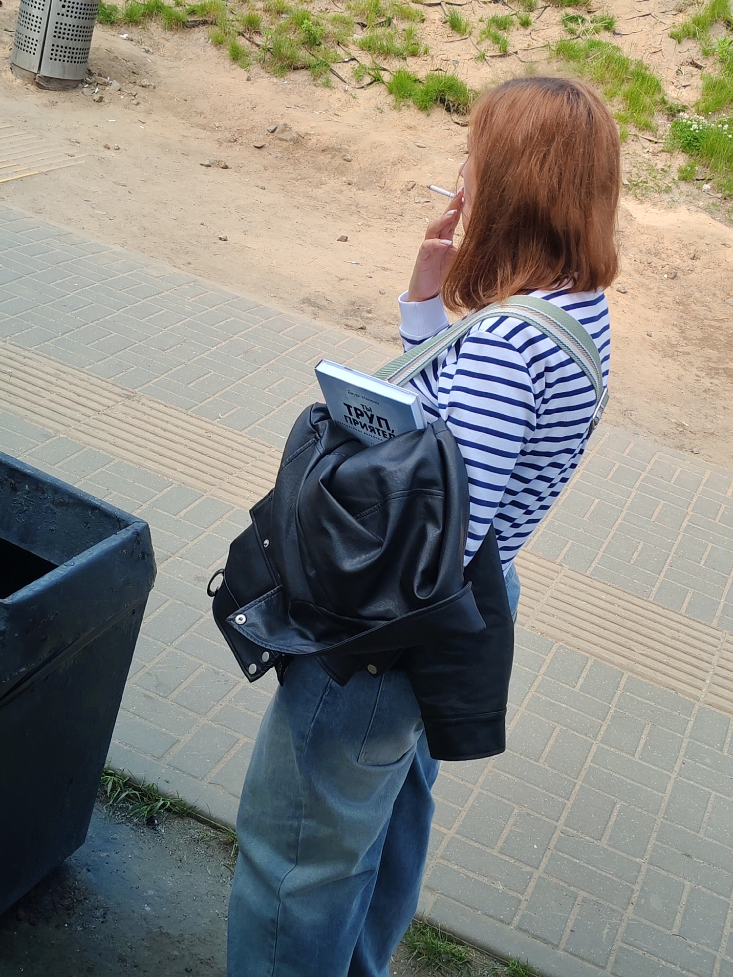The image shows a person, likely female, standing outdoors, viewed from the back and slightly to the side. The individual has shoulder-length reddish-brown hair and is wearing a blue and white striped long-sleeved shirt with blue jeans. They are holding a lit cigarette to their mouth with their right hand, appearing to be smoking.
A light-colored canvas shoulder bag with a striped strap is worn across their body, over which a black leather jacket is casually draped. Tucked into the bag is a light-colored book. Visible text on the book's cover and spine is in Russian, reading "ТЫ ТРУП ПРИЯТЕЛ" (TY TRUP PRIYATEL'), which translates to "YOU ARE A CORPSE, FRIEND." The author's name, "Дилан М" (Dylan M), is also partially visible.
The person is standing on grey, rectangular paved stones, which include tactile strips. To their left is a dark, rectangular trash bin, and further left, a metallic, cylindrical, perforated trash can is partially visible. Beyond the paved area is a sandy, dirt ground with patches of green grass. The bright lighting suggests it is daytime. The scene depicts a casual moment, possibly a break, in an outdoor, urban or semi-urban setting.