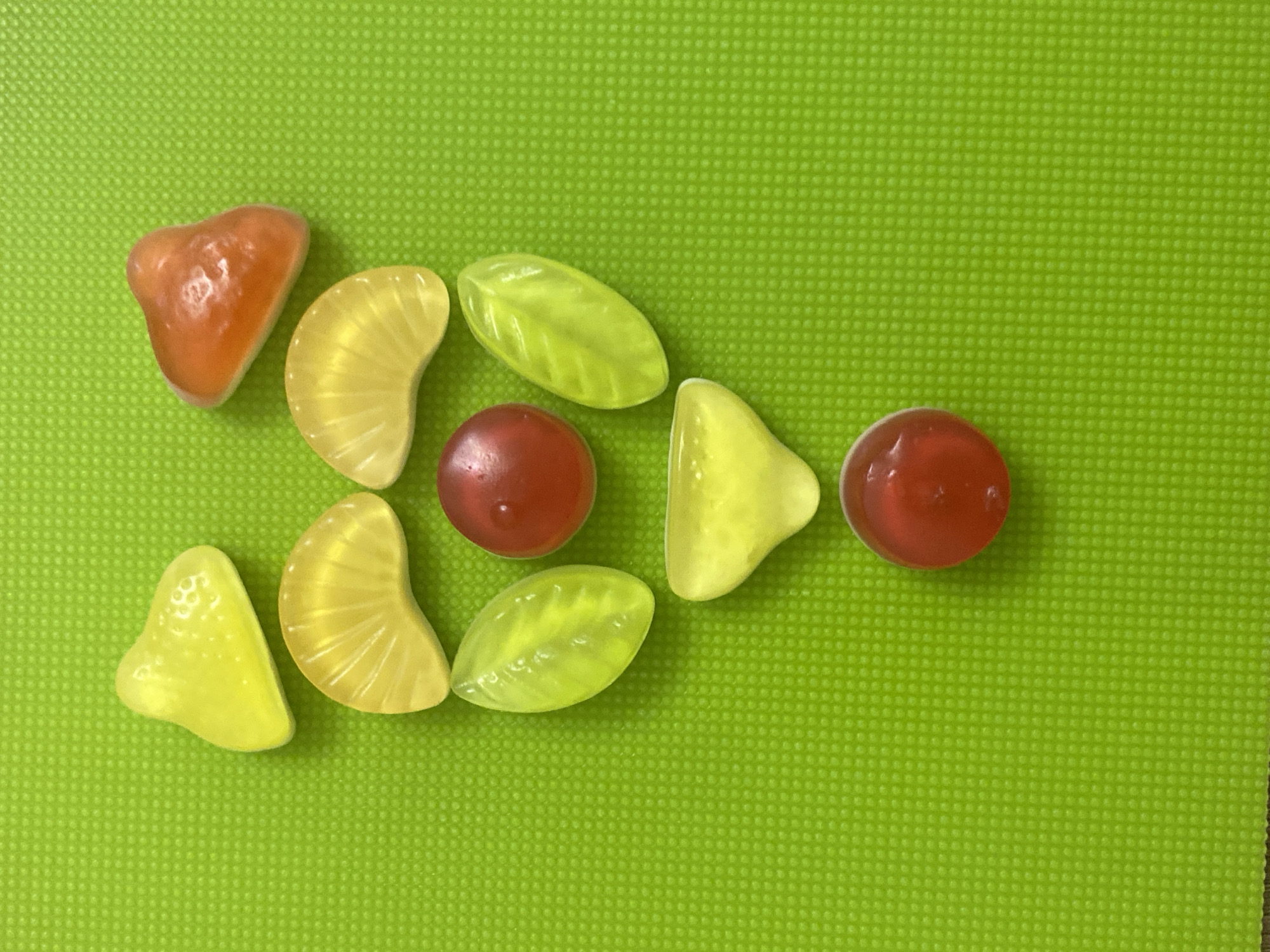 The image displays a collection of colorful gummy candies arranged on a bright green, textured surface. The scene is shot from a slightly overhead perspective, showcasing the candies spread across the frame.
The main subjects are ten translucent gummy candies of varying shapes and colors. There are two red, spherical candies, one near the center and another on the right edge. Three yellow candies are shaped like orange slices, two on the left side and one towards the center-right. Three other yellow candies resemble strawberries or irregular fruit shapes, located on the far left, lower left, and upper right. Completing the assortment are two light green, leaf-shaped candies positioned in the middle, one above and one below the central red candy. The candies appear glossy and slightly reflective.
The background is a uniform, vibrant lime green surface with a fine, repetitive dotted or grid-like texture, suggesting a mat or placemat. The candies are loosely arranged in a curved line or cluster, without any specific activity taking place other than their arrangement for display.
There are no people or specific events depicted. The time of day and weather are not discernible, but the lighting suggests an indoor setting with artificial light. No location cues are visible within the image itself; however, the context provided states the image is from Saint Petersburg, Russia. There is no visible text in the media file.