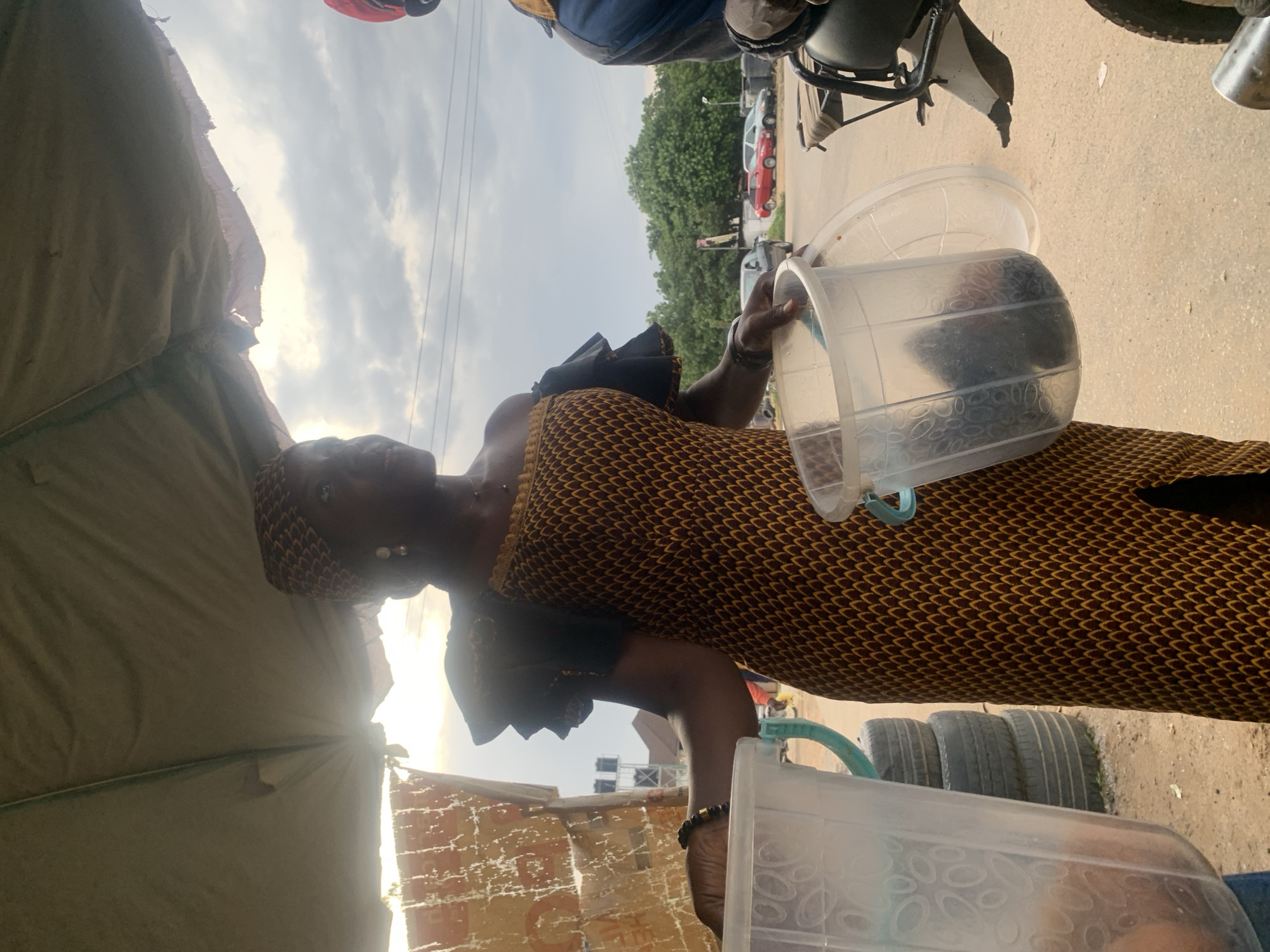 A woman stands under a tan awning, holding a clear plastic container filled with dark objects. She is wearing a patterned dress with off-the-shoulder black ruffled sleeves and a matching headscarf, adorned with dangling earrings. Her right hand, adorned with a black beaded bracelet, rests on the rim of the container in front of her. To her left, a large clear container with a blue handle is partially visible, as is a stack of tires. In the background, a person in a blue jacket and red headwear is seated on a motorcycle, and cars are visible on a street lined with trees. The scene appears to be an outdoor market stall in Abuja, Nigeria, indicated by the style of the awning, the clothing, and the general atmosphere. The lighting suggests it is daytime, possibly late afternoon, with clouds present in the sky.