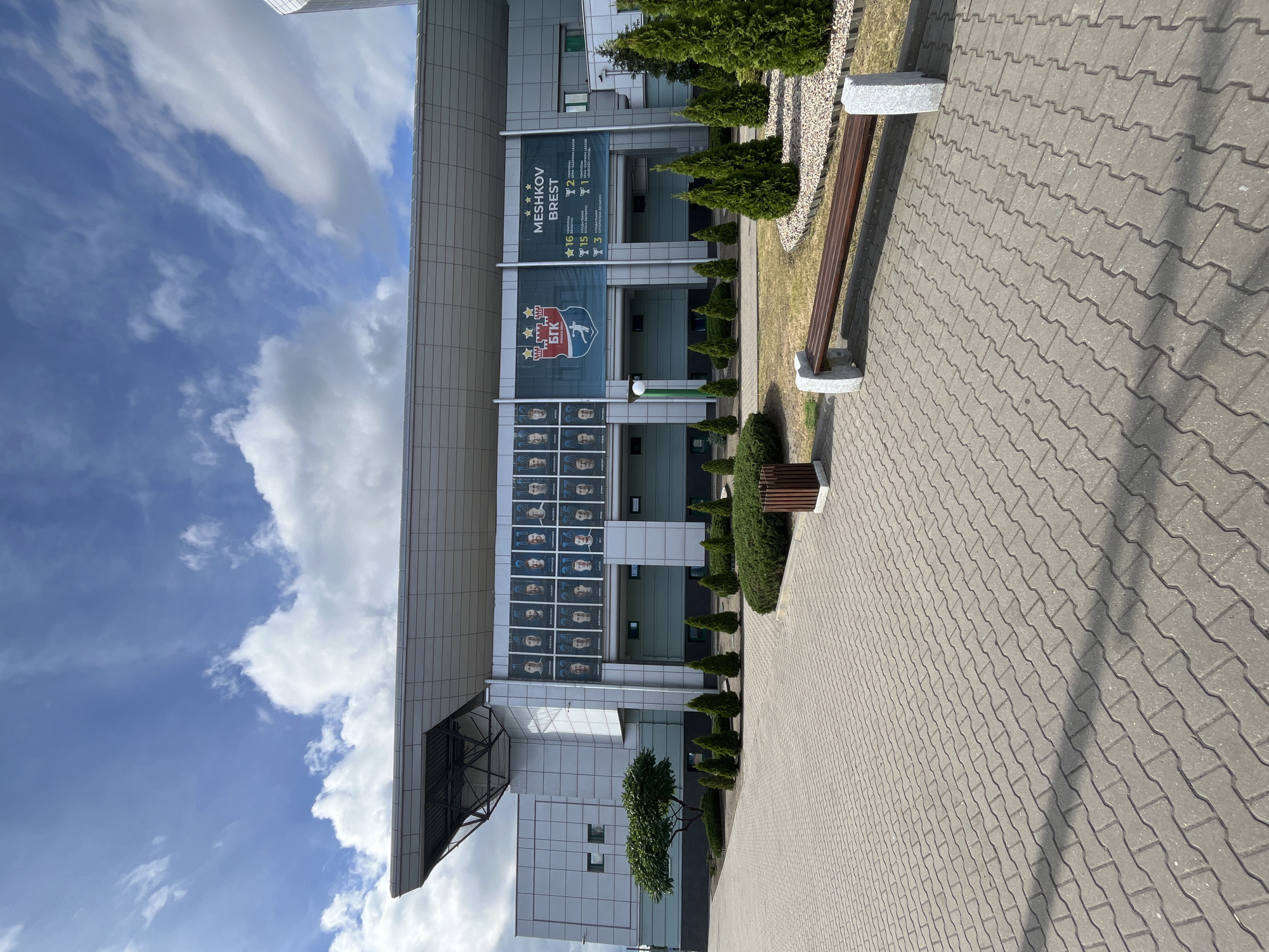 This is a wide-angle, eye-level shot of the exterior of a modern sports complex or stadium, with a blue sky and scattered white clouds above. The building features a large, angular roof overhang and a facade adorned with banners and framed portraits. One banner displays the logo of a sports team, featuring stars and a shield, along with the text "BGK". Another banner, to the right, reads "MESHKOV BREST" and lists athletic achievements with numbers and trophy icons. A series of framed portraits of athletes are mounted in a grid on the upper portion of the building.
In the foreground, a paved walkway made of interlocking gray pavers extends across the scene, casting shadows from an unseen light source. To the right of the walkway, a wooden bench with granite supports sits on a patch of dry grass and gravel. Several small, conical evergreen trees are planted in a line along the edge of the walkway, bordering the building's green-trimmed lower facade. A dark-colored trash receptacle is also visible on the right side of the paved area. The overall environment appears to be an outdoor area associated with a sports facility.