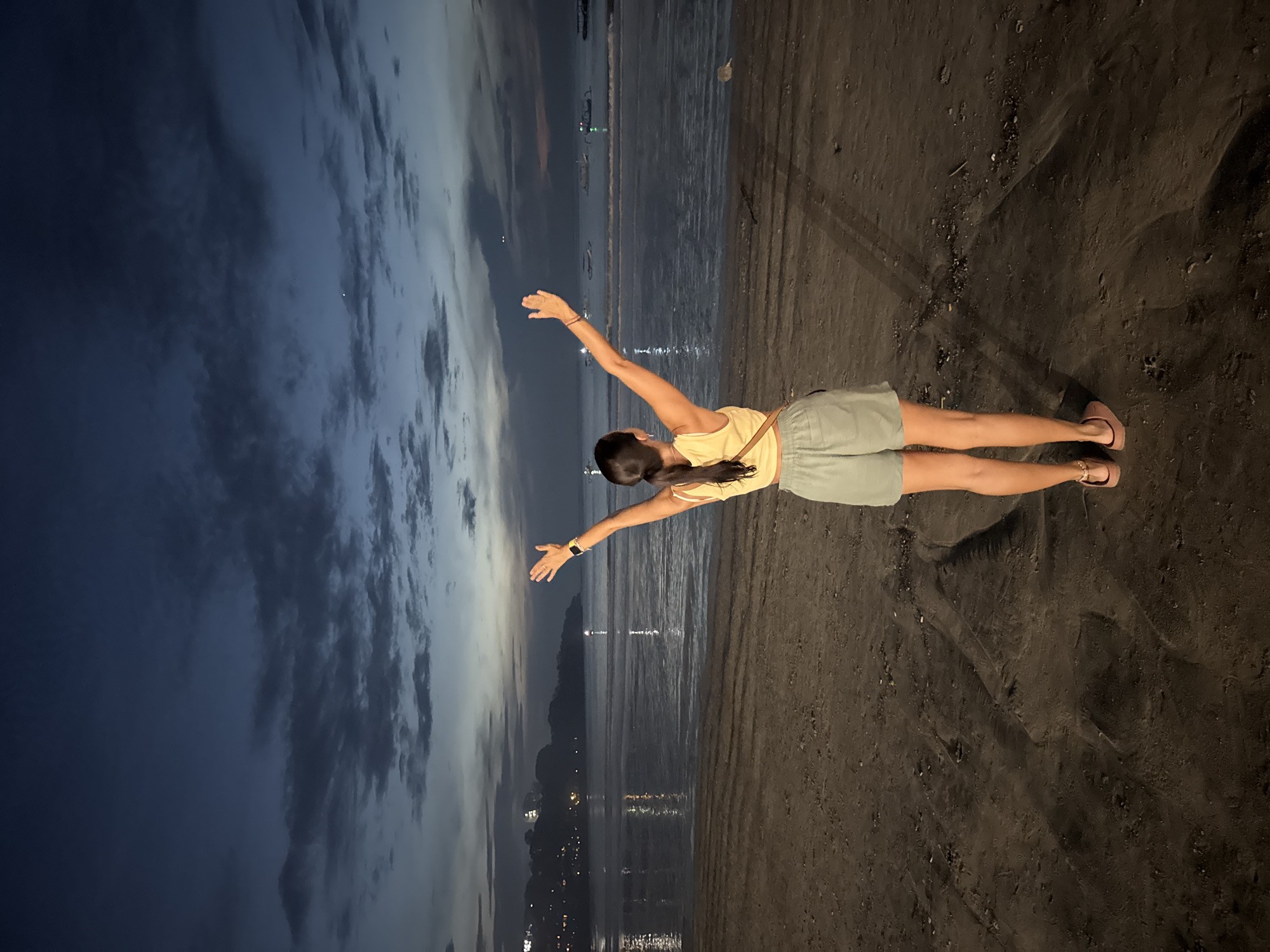 The image shows a woman standing on a dark sandy beach at dusk or night, facing a body of water in Kuta Selatan, Indonesia. She is viewed from behind, with her dark hair in a ponytail, wearing a light yellow crop top, light green shorts, and sandals. Her arms are outstretched horizontally to her sides, suggesting a pose of embracing the surroundings or freedom. A smartwatch is visible on her left wrist.
The foreground consists of dark, wet sand with faint marks. Beyond the woman, the ocean stretches out, reflecting numerous bright artificial lights from a distant coastline, likely a town or city. The water surface is relatively calm, with the light reflections forming shimmering vertical lines. The sky above is dark blue-grey, partially covered by clouds, indicating the late hours of the day. No discernible text is visible in the image. The scene conveys a serene and expansive atmosphere.