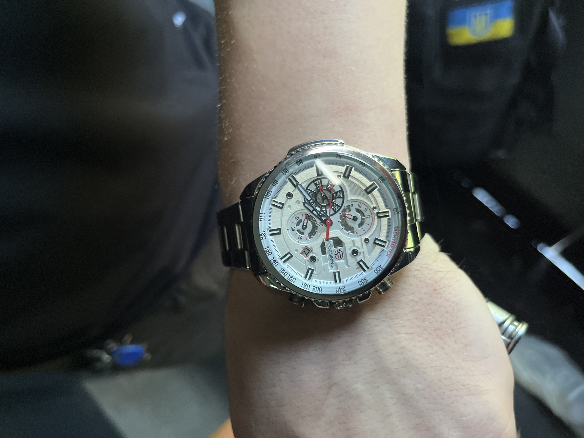 A close-up shot shows a person's wrist wearing a silver and white chronograph watch with a metallic band. The watch face displays multiple sub-dials, Roman numerals, and tachymeter markings. The background is blurred, but a dark fabric garment is visible, along with a hint of a blue keychain. The scene appears to be indoors, possibly in the city of Sviatopetrivske, Ukraine, suggested by a partial view of a blue and yellow Ukrainian symbol in the lower right corner of the frame.
