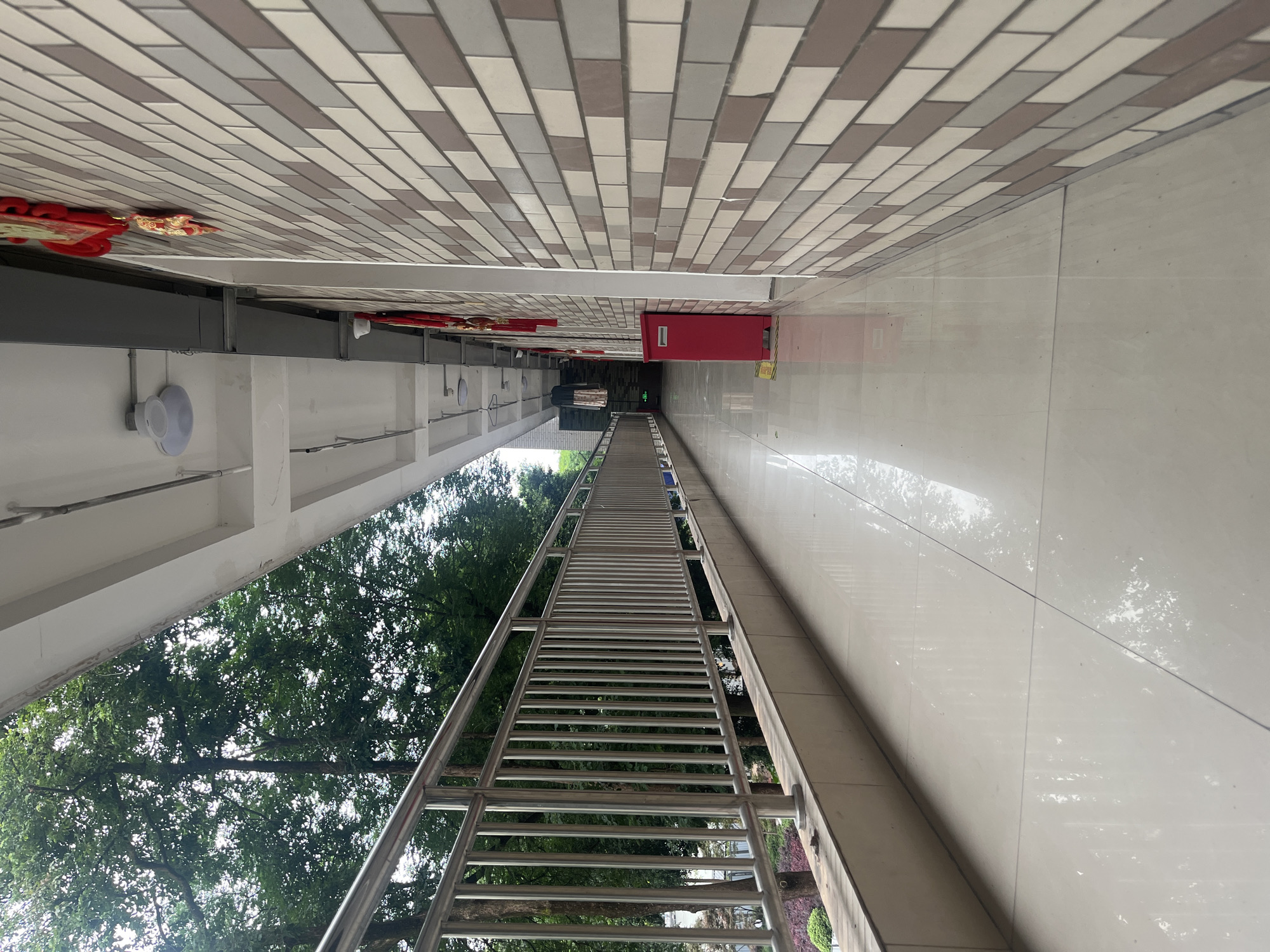This media file captures a long, covered outdoor corridor or elevated walkway, likely part of an institutional or public building in Yuzhong District, China. The perspective emphasizes the length of the passage, receding into a vanishing point in the distance.
The ceiling features a patterned design with alternating light and dark rectangular tiles. Along the left wall, white paint contrasts with several red decorative elements, possibly traditional Chinese knots or festive ornaments, suspended from structural beams. White circular light fixtures or speakers are also mounted on this wall.
The floor on the right is made of glossy, light-colored tiles, reflecting the metal structure and the green trees beyond. A prominent feature is a long, inclined metal structure, resembling a pedestrian bridge or ramp, with numerous horizontal rungs and handrails, extending from the foreground towards the far end. This structure runs alongside the walkway, providing a view of lush green trees that signify an adjacent outdoor area or courtyard.
In the distance, the corridor narrows, leading to a dark opening that suggests another section of the building or an entrance. A bright red cabinet or box is mounted on the right wall near the end of the corridor. A yellow sign is faintly visible on the floor near the red cabinet, but any text on it is unreadable due to the distance and resolution.
The overall scene is well-lit, indicating daytime, and the clean, modern architecture, combined with the traditional red decorations, suggests a blend of functional design and cultural aesthetics commonly found in China. No specific activity or people are present in the image.
