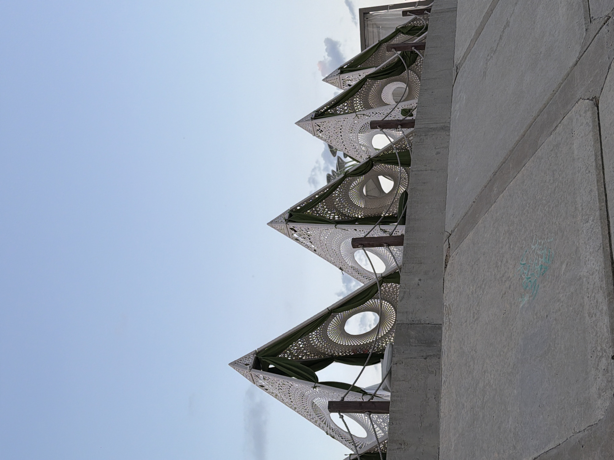 In Fontanka, Ukraine, an outdoor scene captures a series of triangular, pyramid-like structures designed with intricate white lattice work, resembling stylized eyes or spiral patterns. Dark green fabric drapes behind the openings of these structures, creating a visual contrast. The structures are supported by wooden posts and a rope barrier runs along the front. They are situated on top of a concrete wall, which overlooks a lower, sloped concrete embankment. The sky above is a pale blue with scattered, wispy clouds, suggesting it is either early morning or late afternoon. The concrete surfaces show some signs of wear and faint graffiti.
