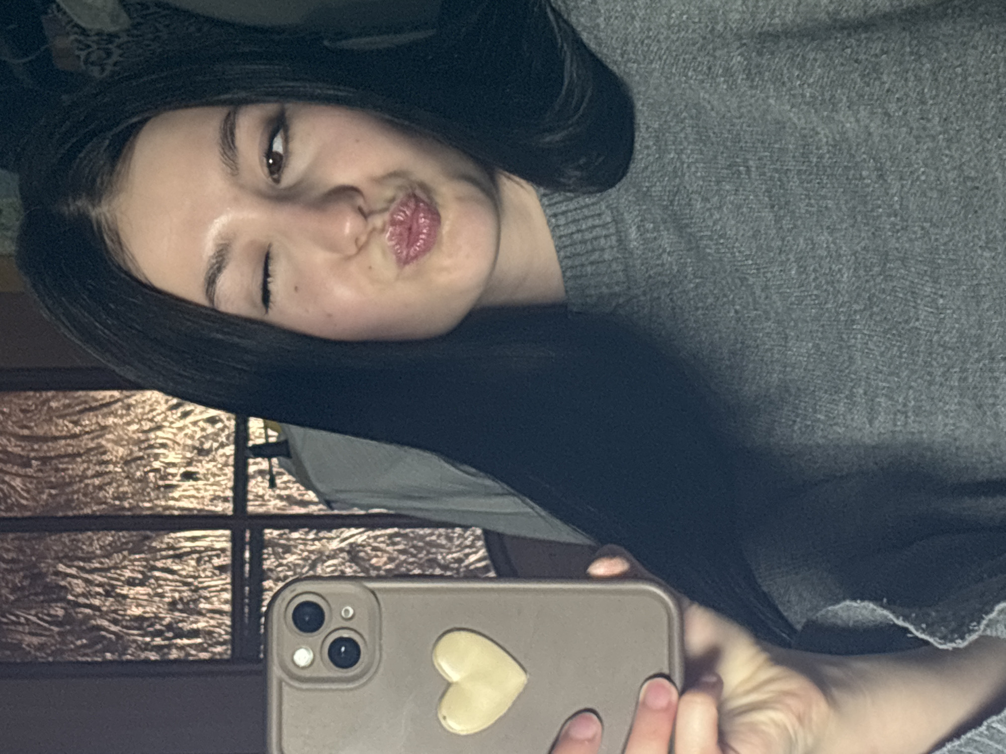 A young woman with long dark hair is taking a selfie. She is winking with her left eye and puckering her lips in a kissing gesture. She is wearing a gray sweater. She is holding a beige iPhone with a heart-shaped sticker on the back. Behind her is a window with multiple panes. The location in the city of Фоки, Russia is not discernible from the image.