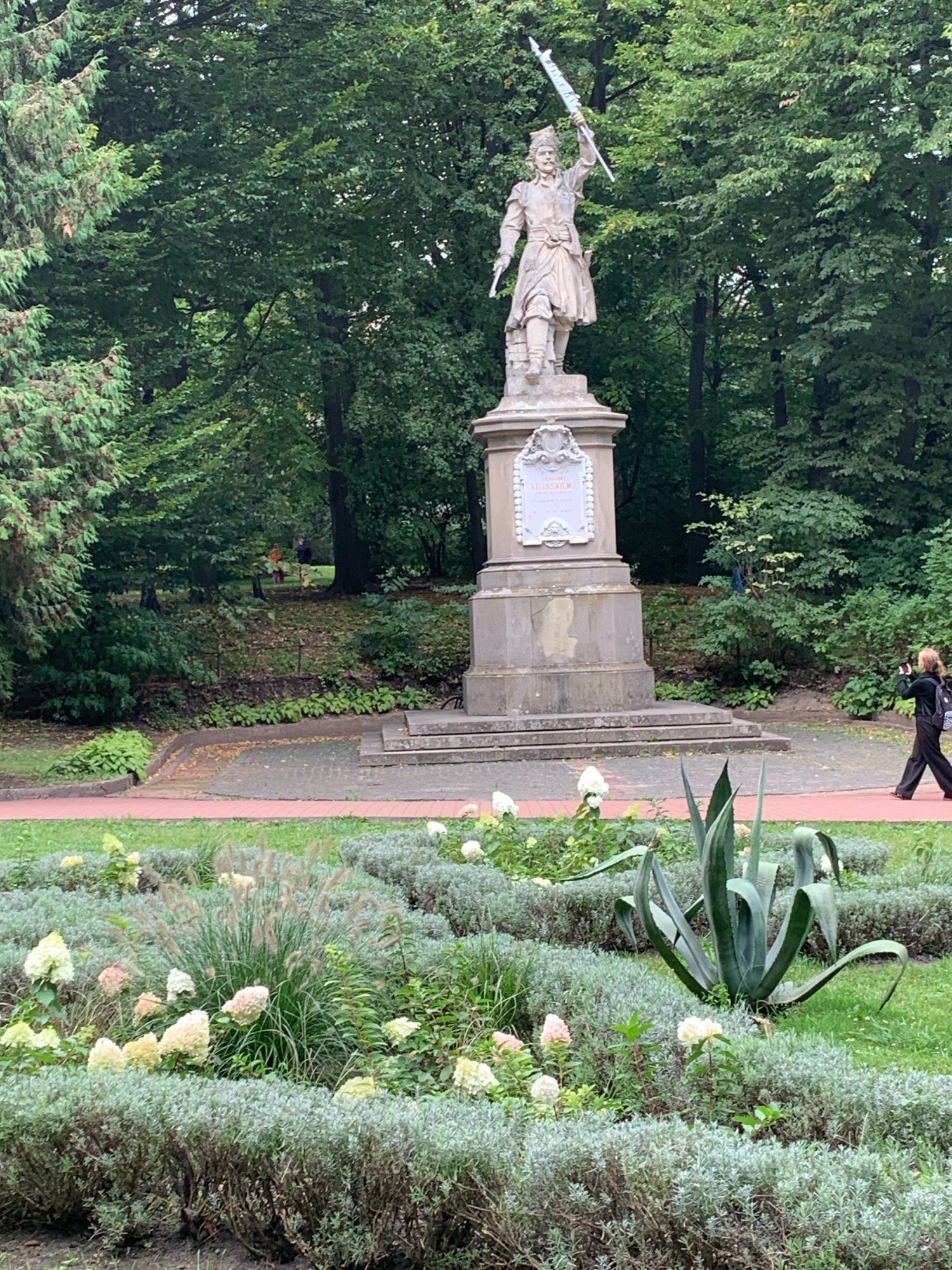 The image depicts a statue of a historical figure in a park setting, with lush green trees and meticulously landscaped gardens in the foreground. The statue stands on a stone pedestal, adorned with a plaque. In the middle ground, a person is walking and taking a photograph with their phone. The foreground is filled with a variety of plants, including tall grasses, hydrangeas, and succulents, arranged in a layered garden design. The park appears to be well-maintained, with paved pathways and manicured hedges. The overall atmosphere is serene and natural.