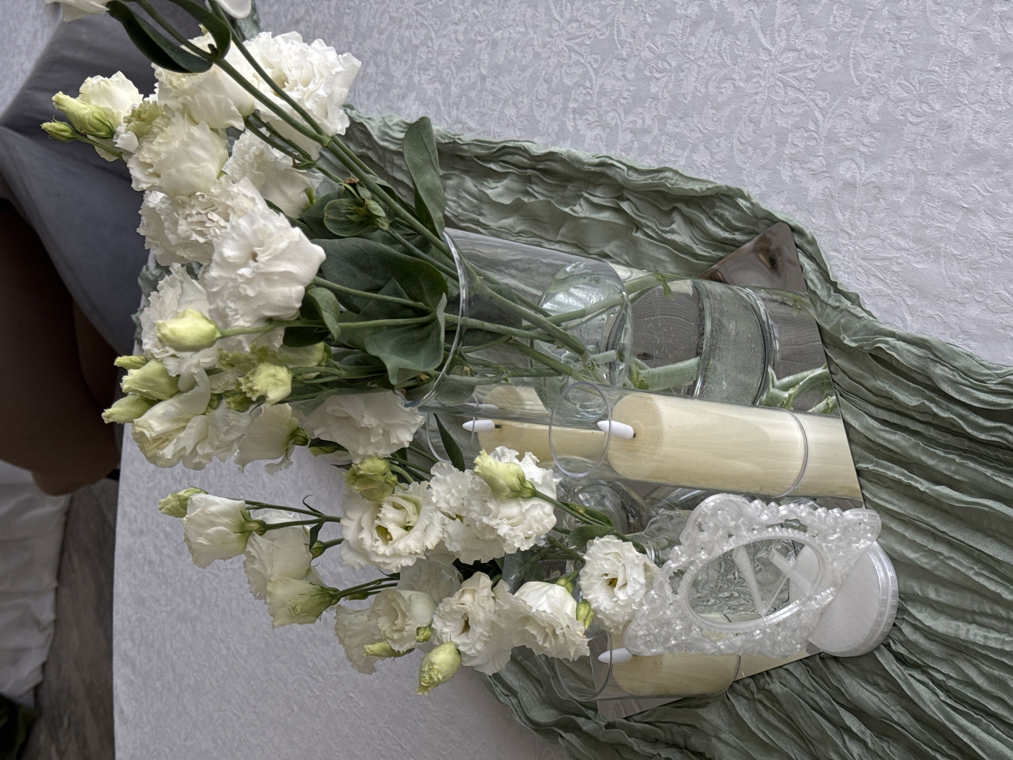 The image depicts a close-up, overhead view of a decorative centerpiece on a white, textured tablecloth. The centerpiece consists of a bouquet of white flowers, likely roses or lisianthus, arranged in a clear glass vase. The flowers have delicate, layered petals and some are in full bloom while others are buds. Surrounding the vase are two tall, cream-colored pillar candles in clear glass cylinders, placed on a mirrored surface. To the left of the candles and vase is a small, ornate, white decorative frame on a white circular base. A textured, sage green fabric runner is draped across the tablecloth, partially obscuring the mirrored surface and acting as a base for the centerpiece elements. The background shows a blurred view of furniture and a person, suggesting an indoor setting. The scene is styled for a table decoration, likely for an event or a home setting. There is no information in the image that would indicate the location is Votkinsk, Russia.