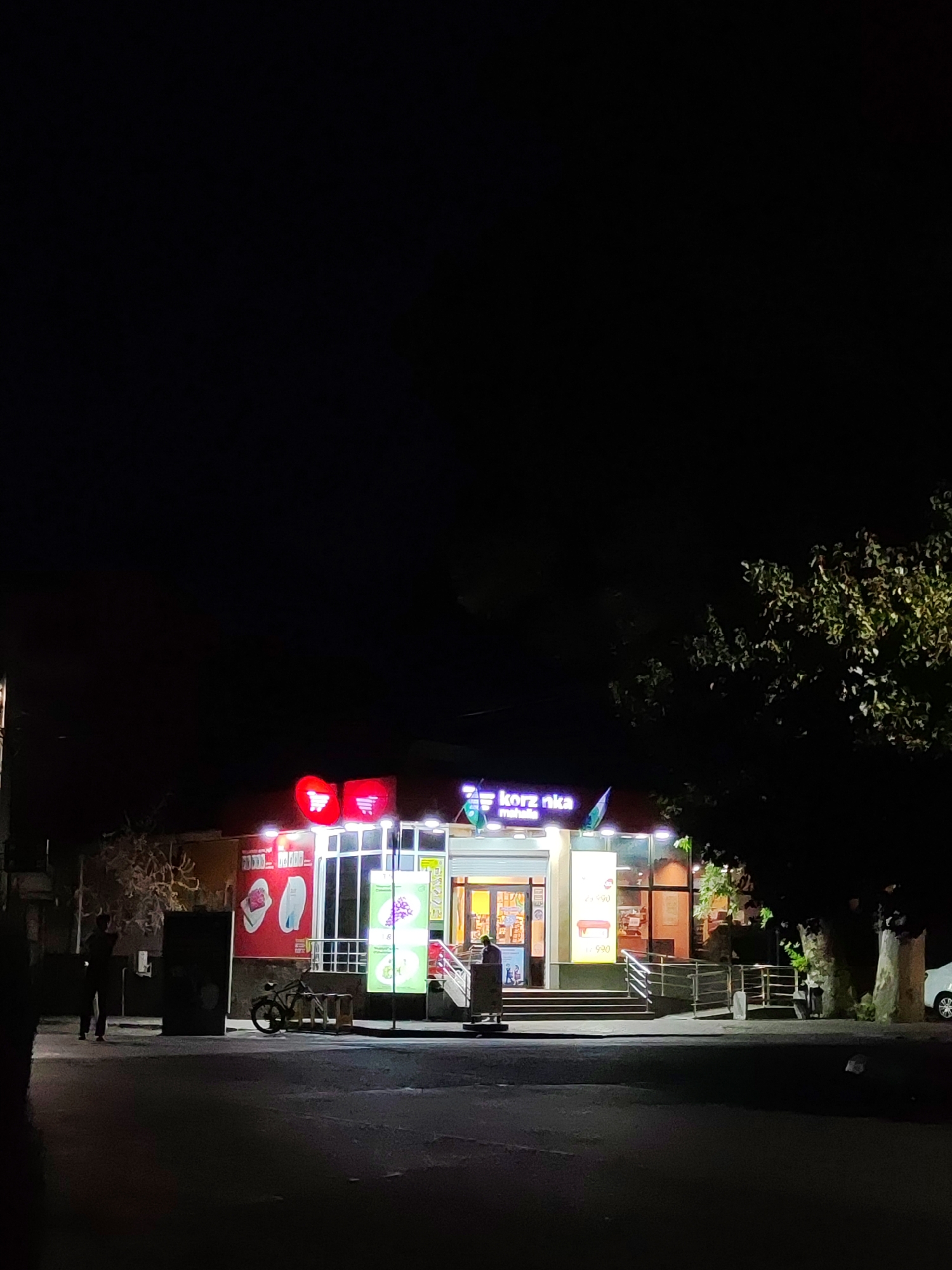 The image depicts a night scene in front of a Korzinka supermarket located in Tashkent, Uzbekistan. The supermarket's facade is illuminated by neon signs, including a red logo with shopping cart icons on the left and the "korz nka" text with a smaller "mahalla" text below it on the right. Several vertical posters advertising products are displayed on the front of the building. To the left of the entrance, a bicycle is parked next to a wooden stand. A person is visible standing to the far left of the frame, near a large speaker system. To the right, a tree with dense foliage partially obscures the view of the supermarket. The area in front of the store is paved, and a ramp leads up to the entrance, which has a set of stairs. A white car is partially visible parked on the right side of the image. The overall atmosphere is dark, with artificial lighting from the supermarket and signs being the primary light sources.