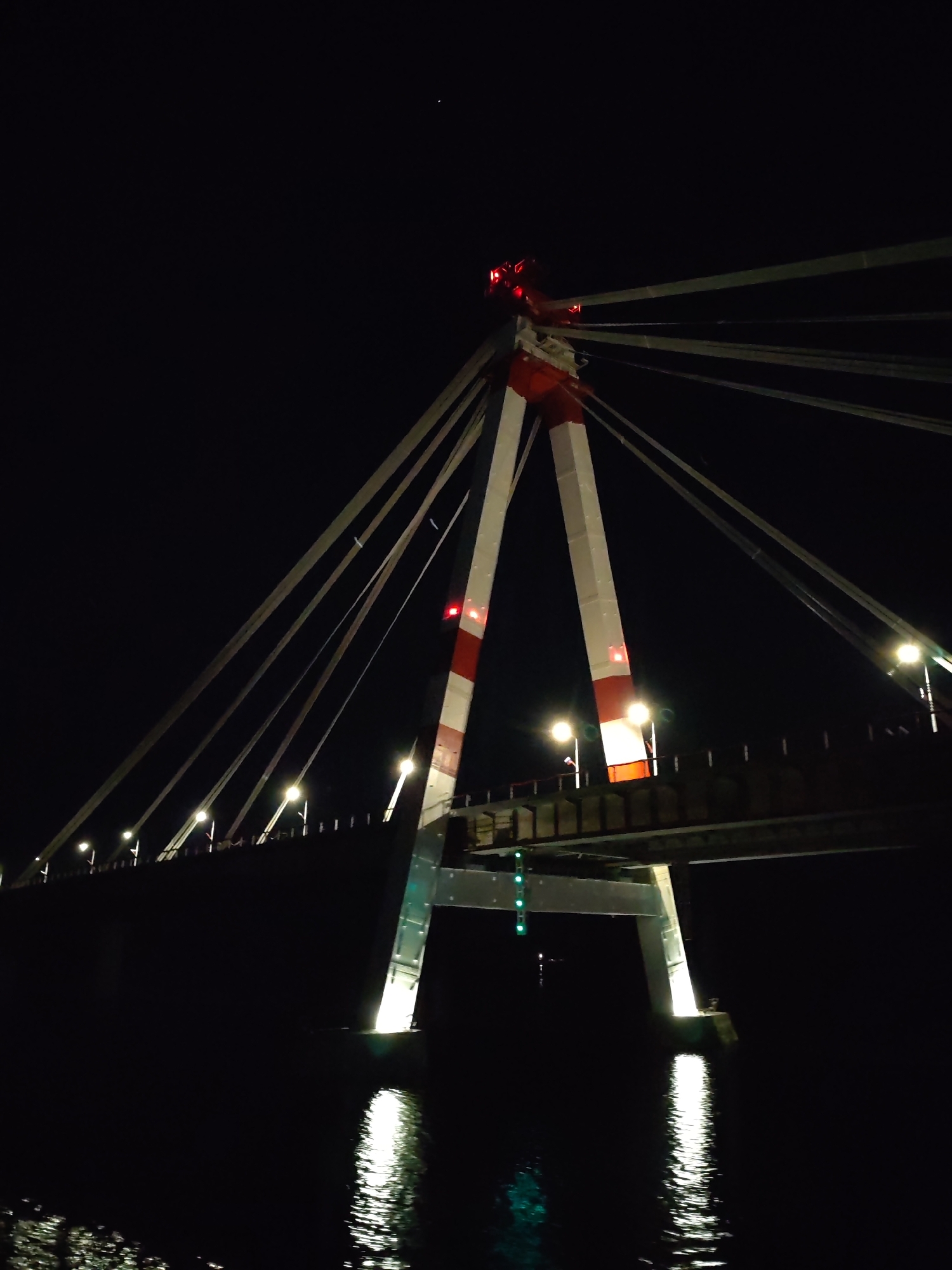 A nighttime view of the cable-stayed bridge in Cherepovets, Russia, is captured. The bridge's central tower, marked with red and white stripes, supports numerous cables extending outwards. Red lights illuminate the top of the tower. The bridge deck is visible, lit by evenly spaced streetlights. The bridge's support structure is reflected in the water below, with green lights visible underneath the bridge. The rest of the scene is dark, suggesting it is night.