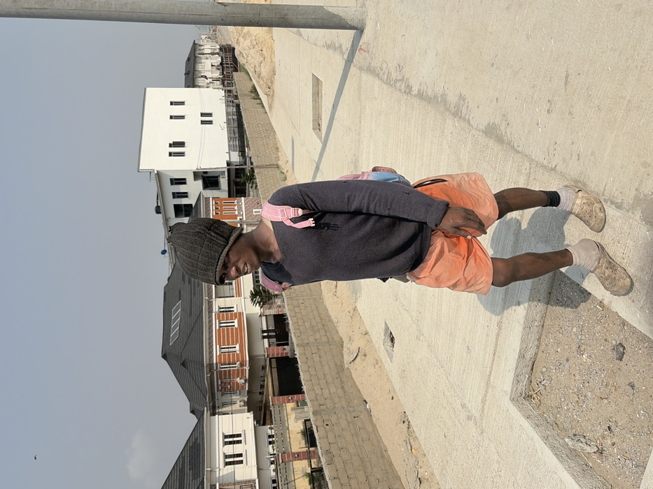A single individual stands facing forward on a concrete path under a bright, hazy sky in Alaguntan, Nigeria. The person, appearing male, is dressed in a dark grey knitted beanie, a long-sleeved dark grey v-neck shirt, orange shorts with a black drawstring, light-colored ankle socks, and light brown, dirt-covered Crocs-style clogs. A pink and light blue backpack is worn on both shoulders. The individual's mouth is slightly ajar, and the gaze is directed towards the left. To the left of the concrete path, an excavated area filled with sand and dirt is visible, bordered by a low cinder block wall. In the background, multiple modern, multi-story residential buildings are present. One prominent house features a dark grey pitched roof with solar panels, white exterior walls, and orange detailing around windows and roofline. Another white building with dark, rectangular windows is visible further right. Shadows indicate bright, direct sunlight. A utility pole stands vertically on the far right of the frame, and a small dark bird is visible in the upper left portion of the sky.