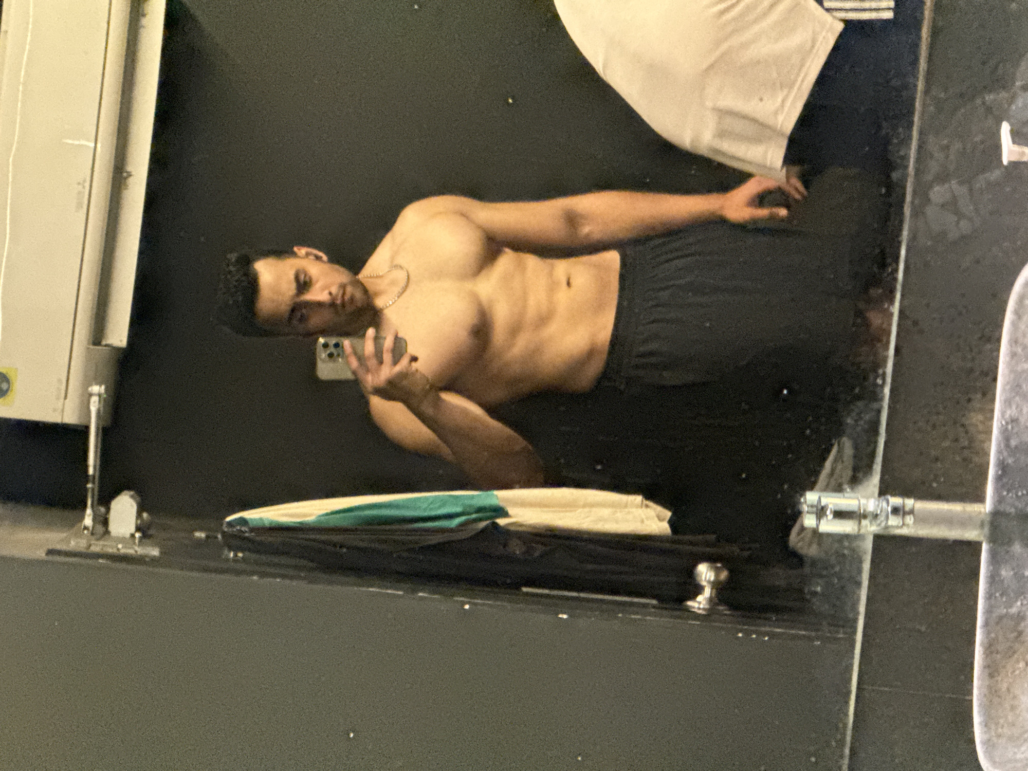 A shirtless man with dark hair and a gold chain is holding a cell phone in a mirror reflection in a gym bathroom, possibly in Kolkata, India. He's wearing black shorts. A partially visible white shirt or towel lies over a black object near the top of the reflection, and the bathroom has a sink and an air conditioner unit.