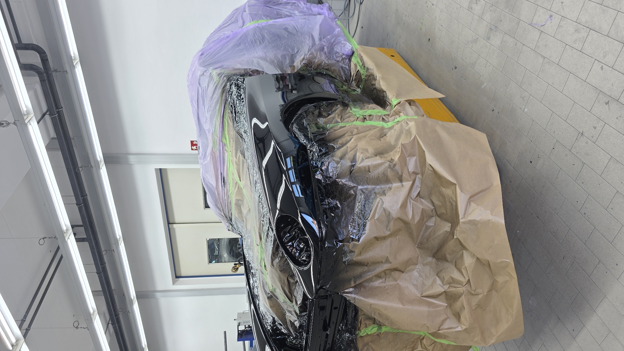 A black sports car is covered in protective plastic and brown paper, presumably for painting or detailing, in what appears to be a workshop or garage. The car is on a yellow platform. The room has fluorescent lighting and white walls. There is a large sliding door in the background.