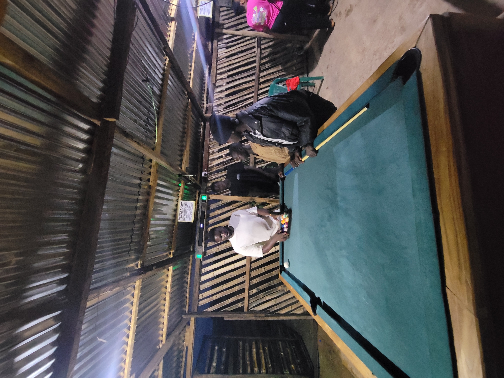 A group of four individuals, three males and one female, are gathered around a pool table in an indoor establishment in Jos, Nigeria. The space has a rustic interior, with a corrugated metal roof supported by wooden beams. The walls are constructed from vertical bamboo poles. The pool table itself is covered in a green felt surface, and a rack of colored pool balls is set up. One man, wearing a white t-shirt, is standing directly over the rack of balls. Another man, wearing a black t-shirt and a beanie, is standing to his left. A third man, wearing a light brown sweater and dark pants, is partially visible behind the man in the white t-shirt. A woman in a pink t-shirt and black pants is standing to the right, further away from the pool table. The lighting appears to be dim, with some overhead lights visible. There is a sign on the back wall that reads "DADIIN KOWA HIDA".