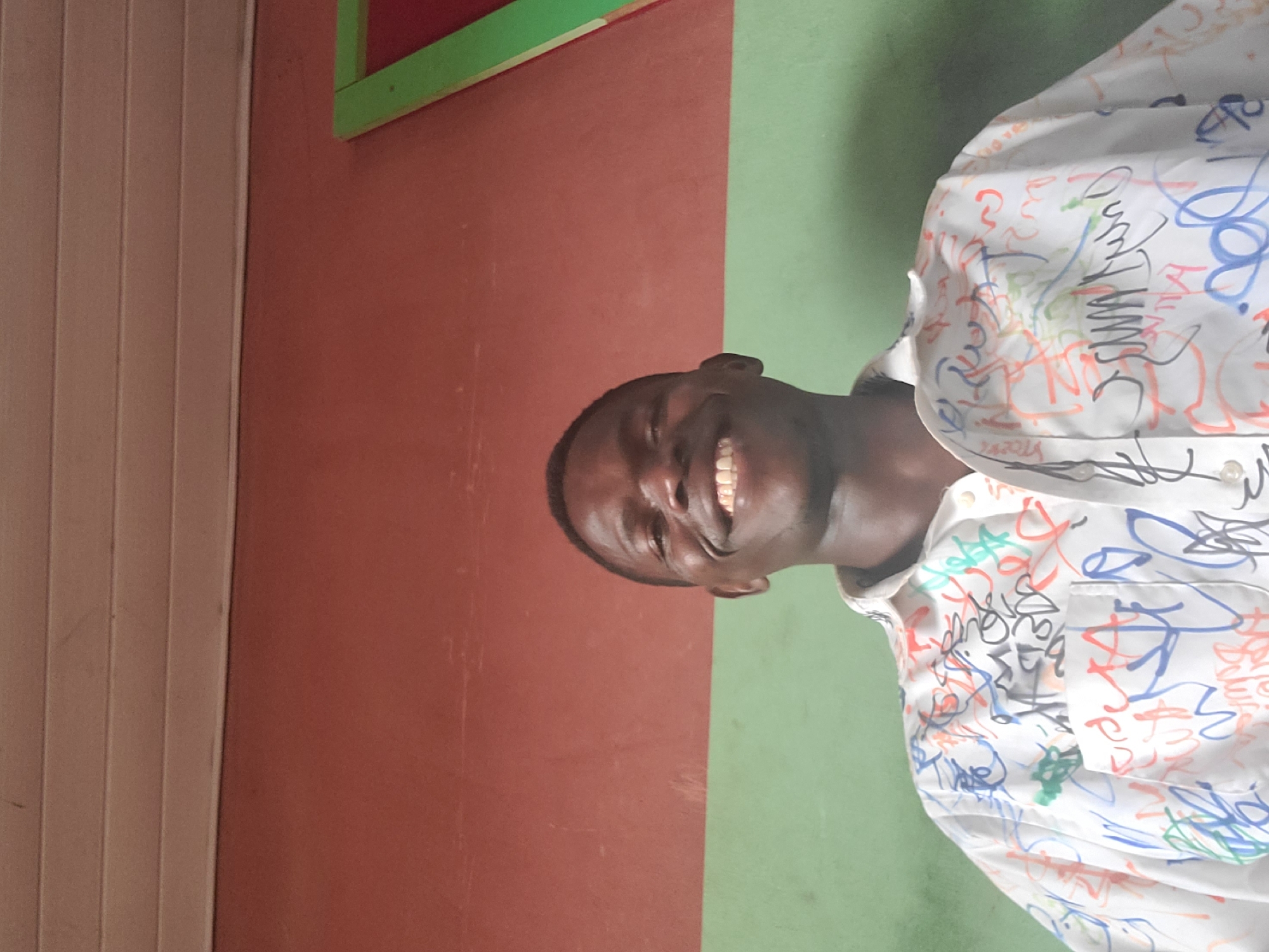 A medium shot captures a smiling Black male in a white button-down shirt adorned with colorful scribbles and writing. He is positioned in front of a wall that is painted in two horizontal sections: red on top and green on the bottom. Above the red section, a cream-colored, slatted ceiling is visible. To the right of the frame, a portion of a window or frame with green and red elements is present. The subject's face is illuminated, highlighting his teeth and the details of the writing on his shirt. The setting appears to be indoors, with the wall and ceiling suggesting a room or a covered outdoor space in Gura Top, Nigeria.