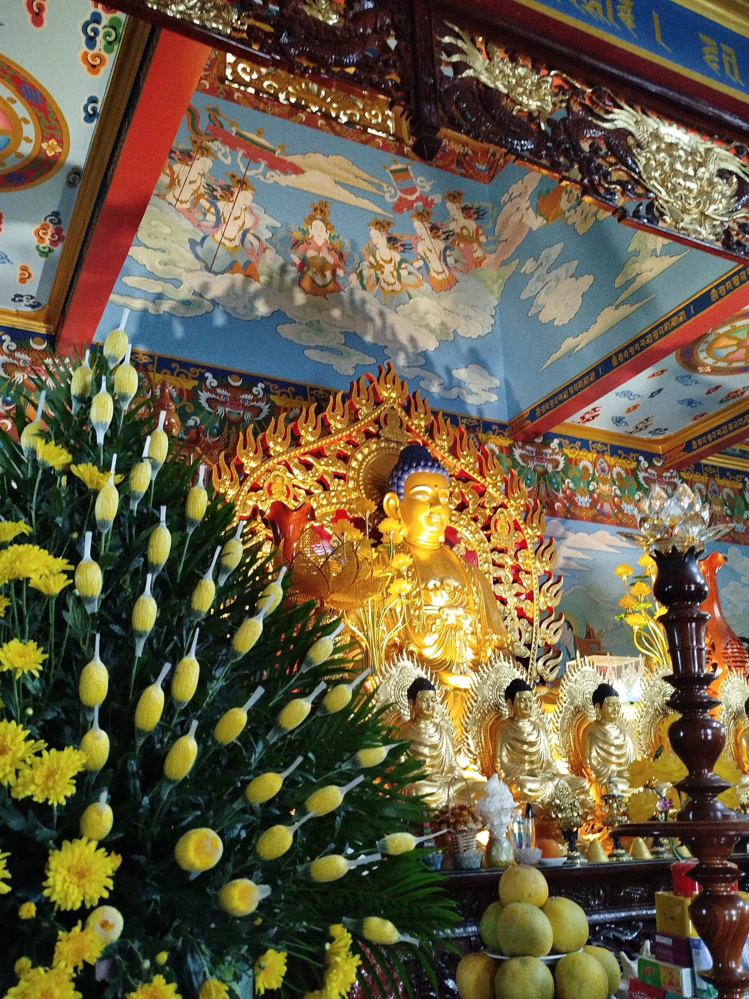 The image depicts the interior of a Buddhist temple, likely in Da Lat, Vietnam, given the artistic style and religious iconography. In the center, a large, golden Buddha statue is prominently displayed, seated with its hands in a meditative gesture. The Buddha wears a serene expression and is adorned with floral garlands. Behind the central statue, a set of smaller golden Buddha statues are arranged, also in seated poses.
The background features ornate, colorful murals painted on the ceiling and walls, depicting celestial beings and mythological scenes in a traditional Asian art style. The colors are vibrant, with blues, reds, yellows, and gold dominating the palette. Intricate gold carvings adorn the upper parts of the walls and the ceiling, adding to the richness of the decor.
In the foreground, to the left, is a large arrangement of yellow flowers and greenery, with numerous yellow, elongated buds or fruits. To the right, a dark wooden altar or pillar is visible, topped with a crystal lotus flower sculpture. A stack of fruits, possibly pomelos, is placed on a lower shelf in front of the altar. Various offerings and decorative items are scattered on the shelves below the Buddha statues, including small ceramic figures and what appear to be books or scrolls. The overall impression is one of reverence and spiritual grandeur.