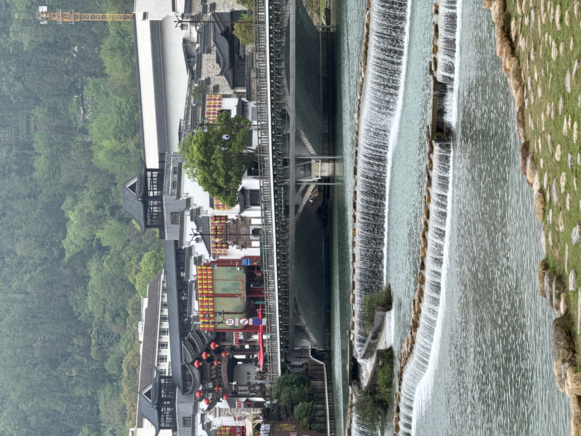 The image displays a picturesque scene of a traditional Chinese-style town situated alongside a distinctive tiered river or stepped waterfall, with a forested mountain backdrop.
In the foreground, a series of artificial or natural cascades creates a multi-level water feature, with water flowing over numerous low stone barriers. The riverbank on the far right is covered in green grass interspersed with light-colored rocks.
The midground features a clear body of water that feeds into these cascades. On the left bank of this water feature, traditional Chinese architecture is prominent, characterized by dark, intricate tiled roofs and white or light-colored walls. Numerous red and yellow lanterns adorn the buildings, suggesting a vibrant cultural atmosphere. A lush green tree stands out among the structures. A large, dark bridge or elevated walkway spans the water, supported by many vertical posts, with what appear to be small boats or structures beneath it.
In the background, dense green forests cover rolling hills or mountains. A tall, yellow construction crane is visible extending above the treeline and buildings in the upper left portion of the image, indicating ongoing development.
The scene is lit by soft, diffused light, suggesting an overcast day. Visible text includes a red "50" speed limit sign and another red prohibition sign located near the bridge structure. Chinese characters are present on some lanterns and building signs, though mostly indistinct. The setting is Wulingyuan, China, and the image captures a blend of traditional aesthetics and modern elements. No specific human activity is the central focus, rather a general landscape view.