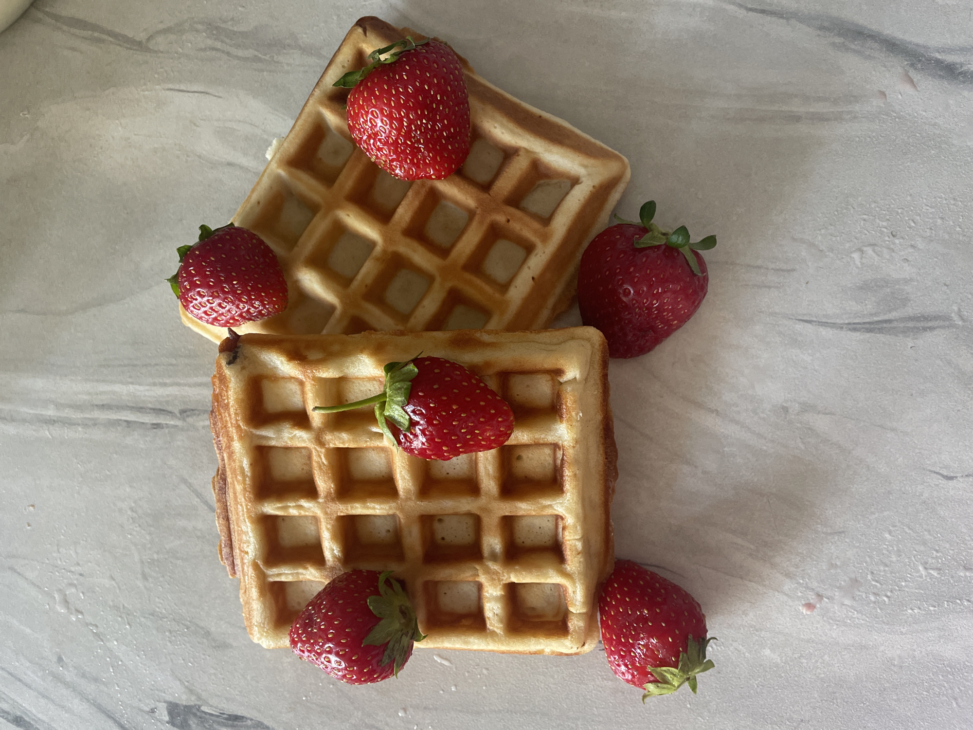 This is a top-down, eye-level, medium shot of two square-shaped waffles, each topped with fresh strawberries, arranged on a light gray, marbled surface. One waffle is positioned diagonally in the upper half of the frame, and the other is placed below it, also at an angle. Five whole strawberries are visible: one atop the upper waffle, one to its right, one on the left side of the lower waffle, and two more to the right and bottom right of the lower waffle. The waffles are golden brown with a distinct grid pattern. The strawberries are bright red with visible seeds and green stems. The marbled surface has subtle gray and white veining, creating a clean and simple background. The lighting is bright and even, suggesting an indoor setting, possibly a kitchen or dining area, illuminated by natural light. There is no text visible in the image. The overall impression is one of a simple, fresh, and appealing breakfast or dessert.