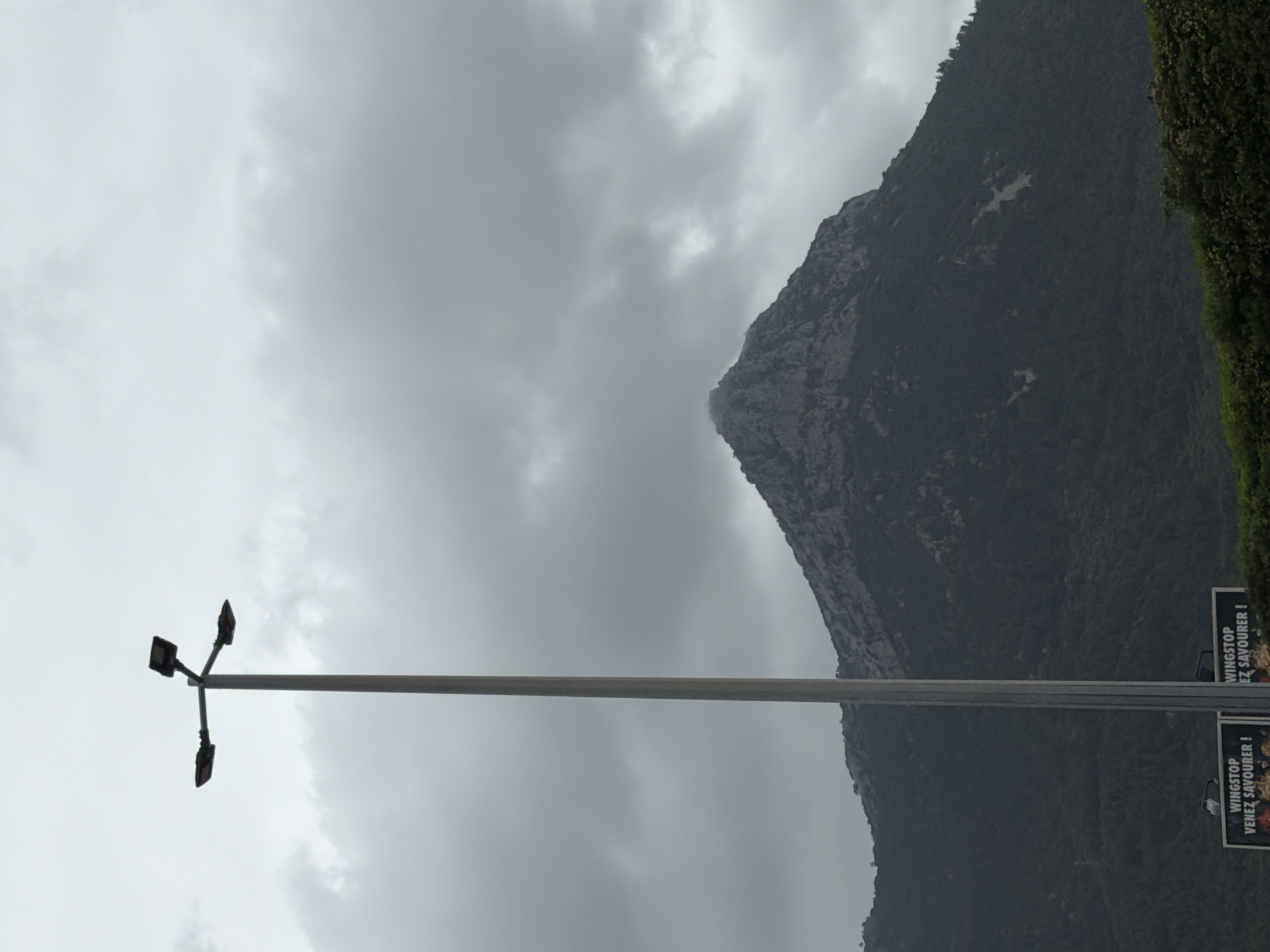 The image shows a vertical view of a cloudy sky over a mountain in La Garde, France. A tall light pole is on the left, and a sign advertising "Wingstop, Venez Savourer!" is on the right. The overall scene is overcast.