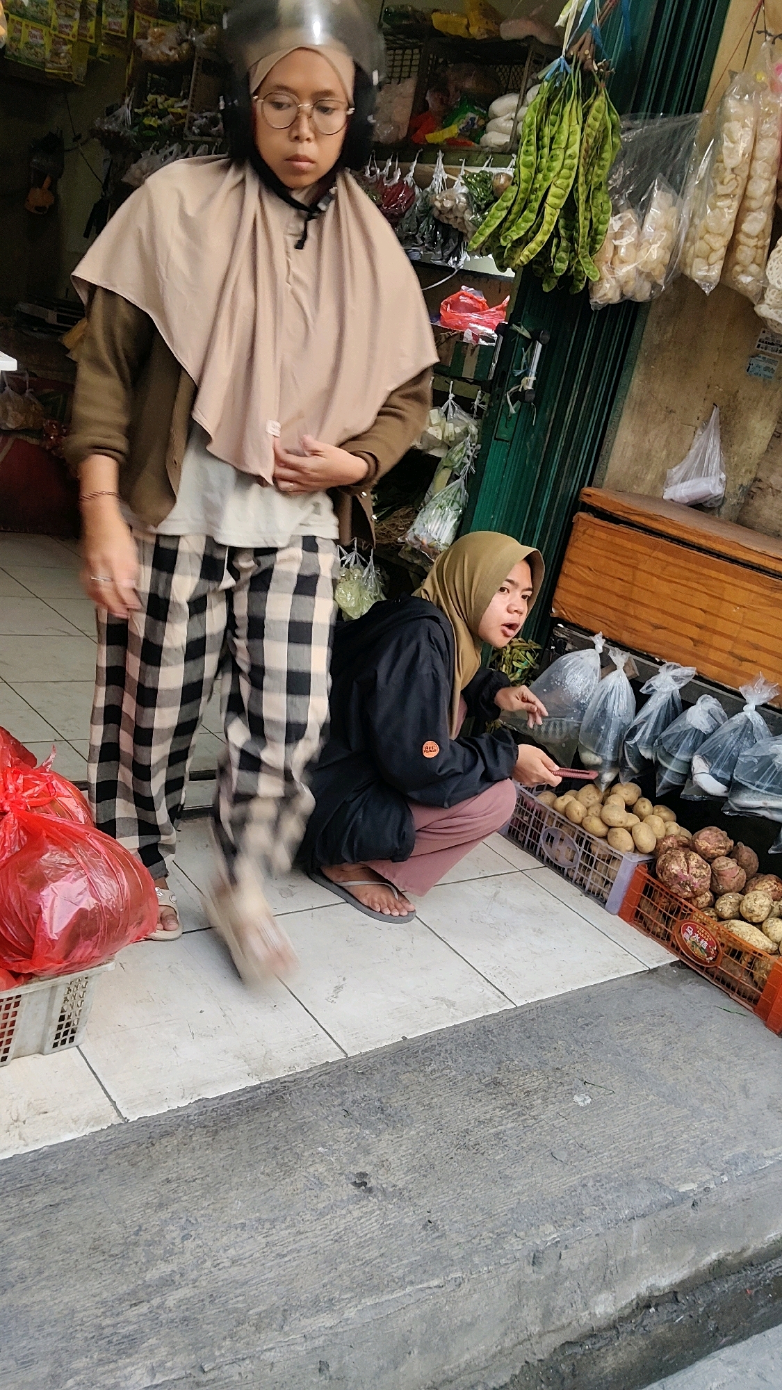 The image captures a street scene outside a market stall in Mekarmukti, Indonesia. In the foreground, a person wearing a helmet and glasses, with a light brown headscarf and a brown jacket over a light-colored shirt, stands with their hands clasped. They are wearing black and white checkered pants and white sandals. To the right, another person wearing a tan hijab and a black jacket over a pink shirt is crouching down, examining produce. This person is wearing grey flip-flops and is holding a red object. In front of them are two crates of vegetables, one filled with potatoes and another with sweet potatoes. Behind the people, the market stall displays various goods, including green beans, packaged snacks, and other produce. The ground is tiled, and a concrete step is visible at the bottom of the frame. A large red plastic bag sits in a white basket to the left.
