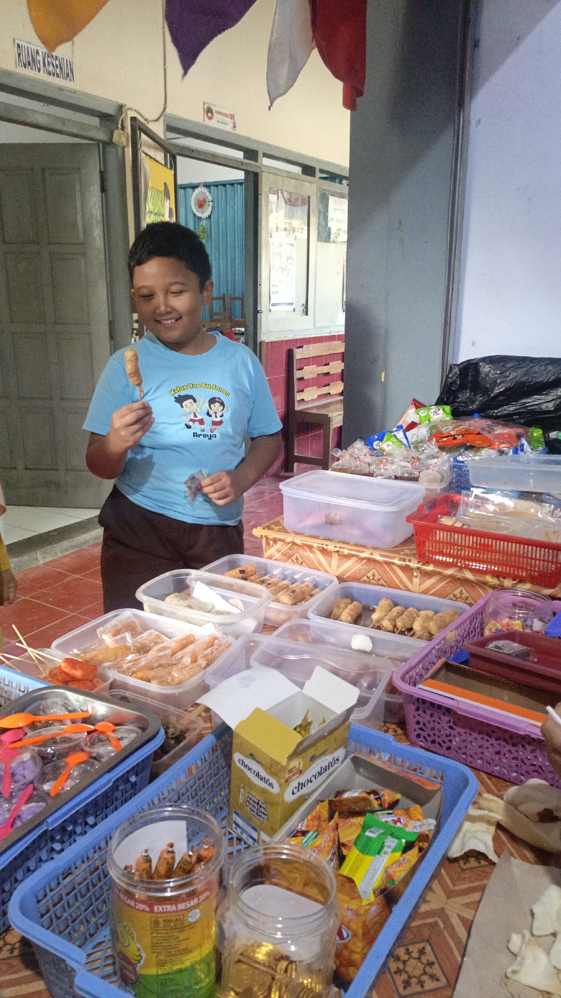 A young boy, likely of Indonesian descent, stands in front of a table laden with various snacks and food items. He is wearing a light blue t-shirt with graphics and text that read "Kelas Yes Budono" and "Arsya." He is holding a skewer with a small, fried food item, appearing to be tasting or about to taste it. His mouth is slightly open, and he is smiling, suggesting enjoyment.
The table is covered with plastic containers filled with an assortment of food. These include what appear to be fried snacks like spring rolls or similar items, some with meat or other fillings, as well as some individually wrapped candies or small snacks. There are also baskets filled with packaged snacks, such as "Chocolatos" brand wafer rolls, and other colorful snack bags. A jar filled with what looks like small, wrapped candies is also visible.
The background reveals an indoor setting, possibly a school or community hall, indicated by the sign "RUANG KESENIAN" (Art Room) above a doorway. Flags or fabric banners in purple, white, and red are hanging from the ceiling. The walls appear to be a light color, and there are doors and possibly windows in the background. The flooring is tiled with a reddish-brown pattern. The overall scene suggests a snack sale or a distribution event. The presence of the "RUANG KESENIAN" sign and the Indonesian text on the boy's shirt point to a location within Indonesia, possibly Salatiga, as implied by the prompt.