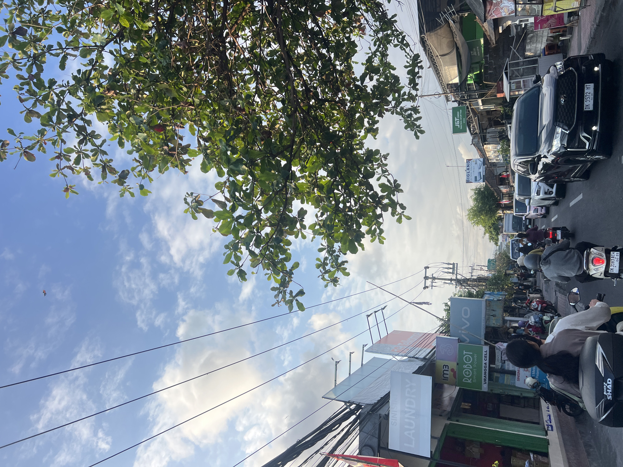 The image captures a street scene in Denpasar, Indonesia, with a focus on a busy road and overhead power lines. In the foreground, two individuals are riding a motorcycle, with the rider in the rear facing away from the camera. They are in traffic alongside other motorcycles and cars. Shops line the right side of the street, with various signs visible, including "SINISKI LAUNDRY," "IMB," "ROBOT Smart Power," and "VIVO." Further down the road, a black SUV with license plate "DK 1572 HT" is prominent in the lane. Overhead, a dense canopy of green leaves from a tree partially obscures the sky. The sky is partly cloudy, with a small kite visible in the upper left quadrant. The overall atmosphere suggests a typical day with moderate traffic on an urban street.