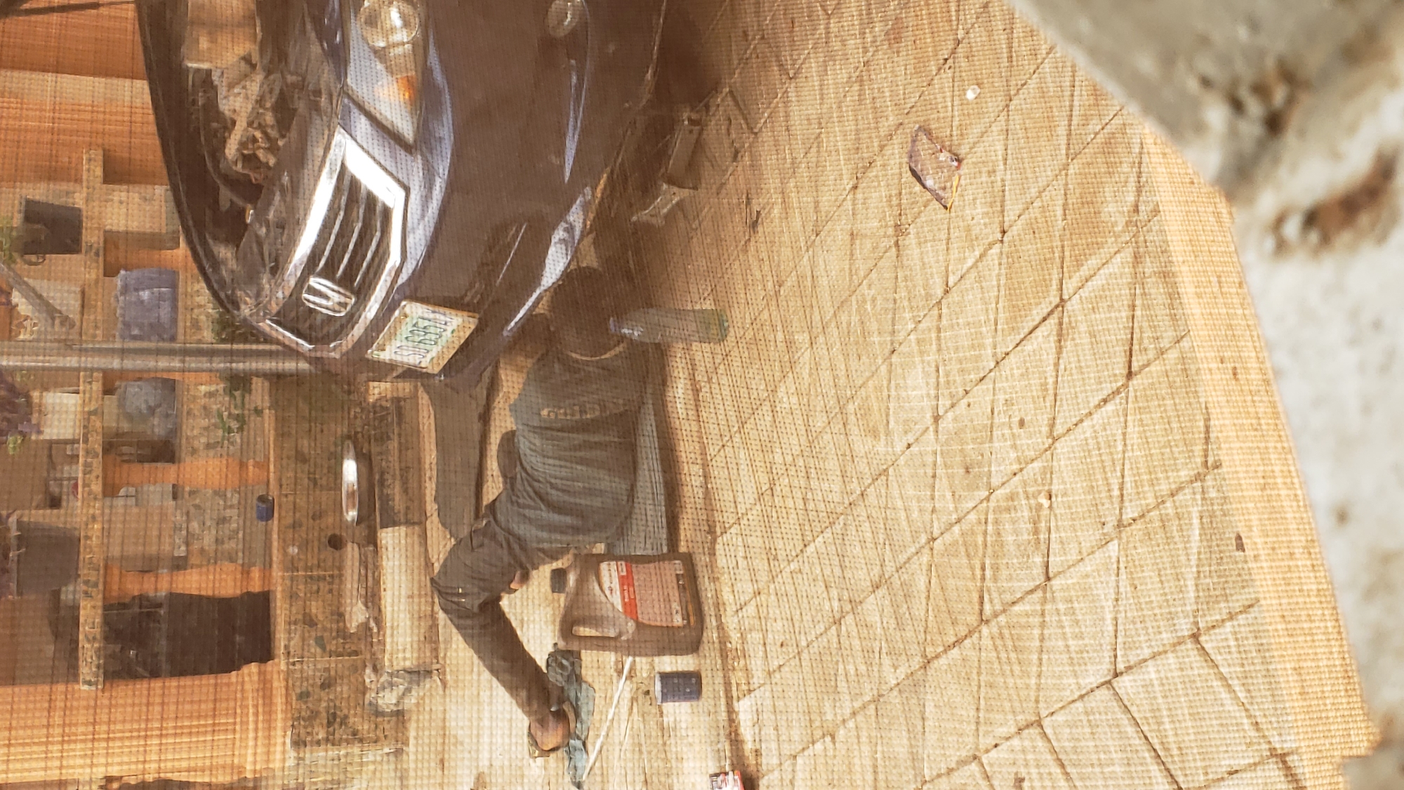 A man is lying on his back under a blue car, appearing to be performing maintenance or repairs. The car's front is visible, including the license plate, grill, and partially open hood. An oil container is near the man on the ground, along with tools and other objects. The setting appears to be outdoors, on a surface with a pattern. Based on the license plate and the prompt's specification, the scene is located in Sabon Tasha, Nigeria.