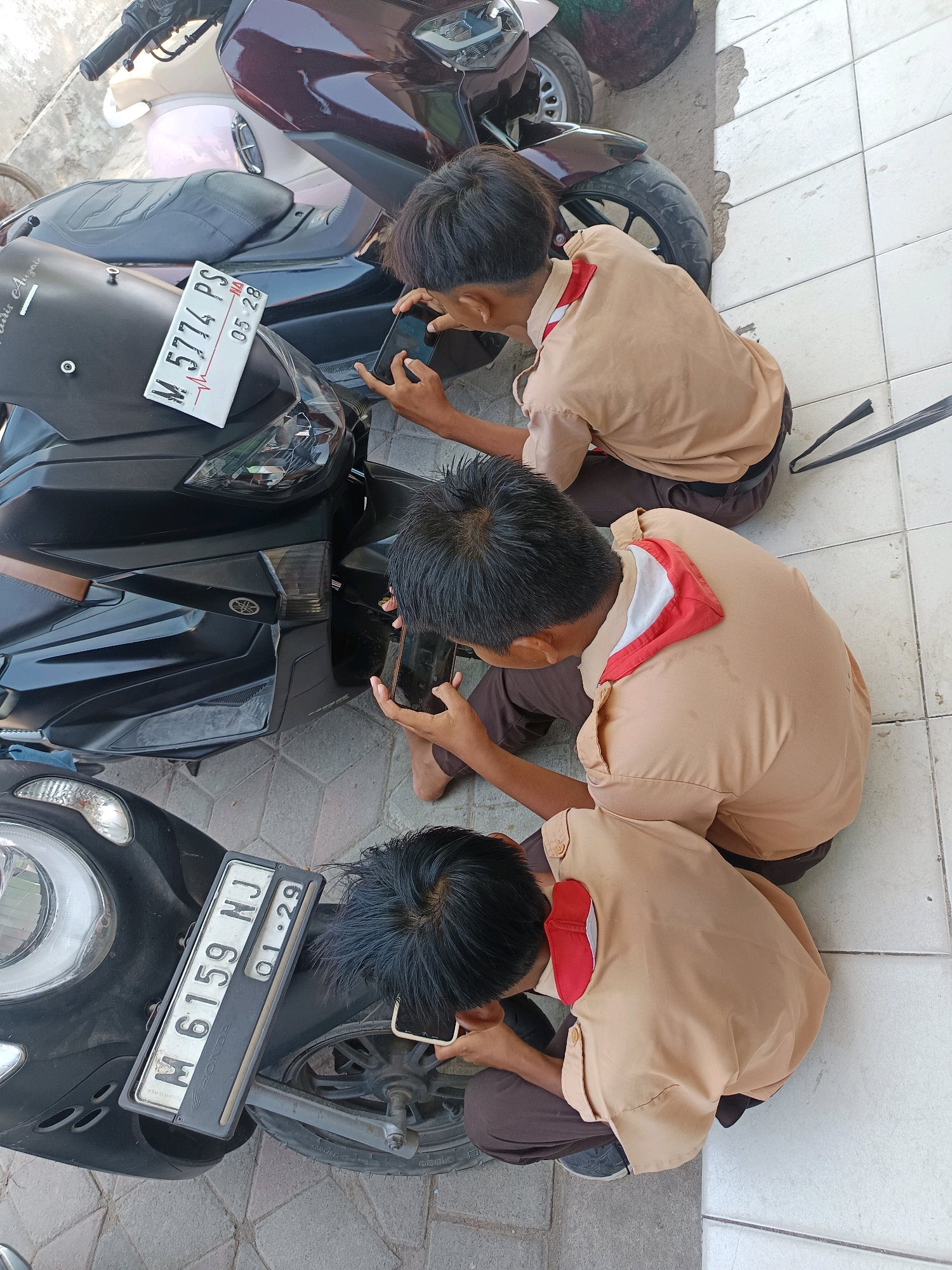 Three young boys wearing light brown uniforms with red and white neckerchiefs are sitting on the ground next to several motorcycles. Two of the boys are looking at their phones. One motorcycle in the foreground has a license plate that reads "M 6159 NJ". Another motorcycle in the background has a license plate that reads "M 5774 PS". The scene appears to be outdoors on a tiled surface next to a paved area. The location is identified as Labuhan, Indonesia, based on the license plate format and common attire for Indonesian schoolchildren.