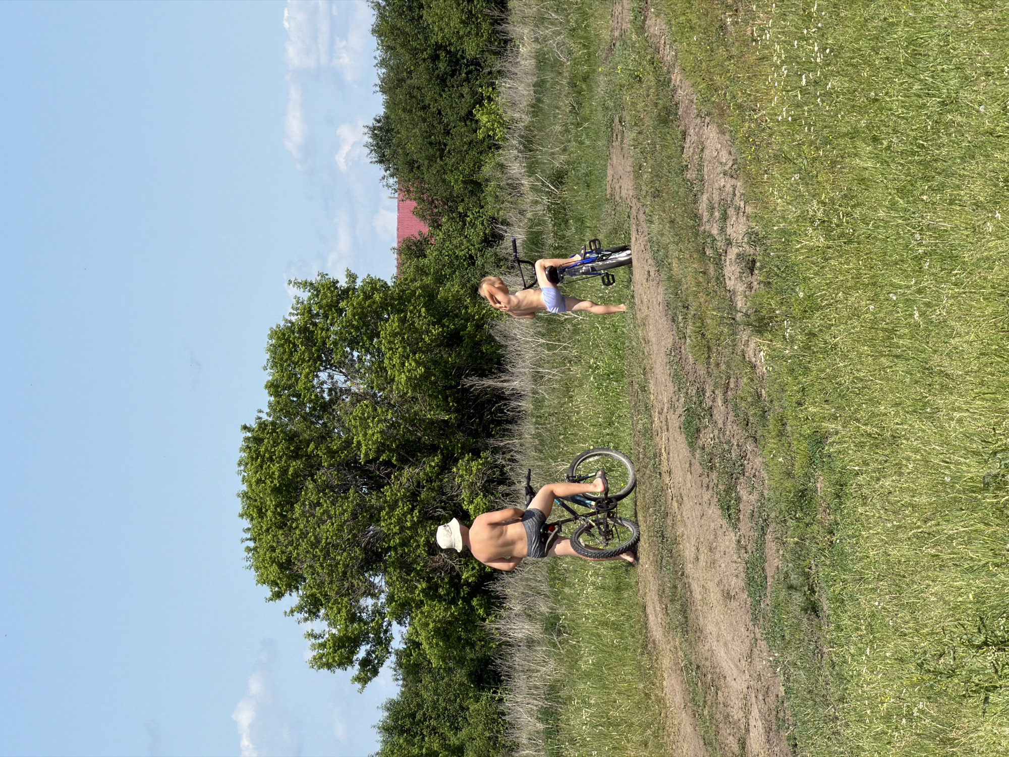 Two individuals are present in an open, grassy field with a dirt path. The person on the left, facing away from the camera, is an adult wearing a bucket hat and shorts, standing with a bicycle. The person on the right, also facing away, is a child wearing light-colored shorts, sitting on a bicycle. Both individuals appear to be shirtless. The background consists of dense green trees, with a glimpse of a red-roofed building visible through the foliage. The sky is clear and blue with a few scattered clouds. The scene is outdoors in a rural or semi-rural setting, likely in or near Zelenyi Dol, Russia, given the context of the question.