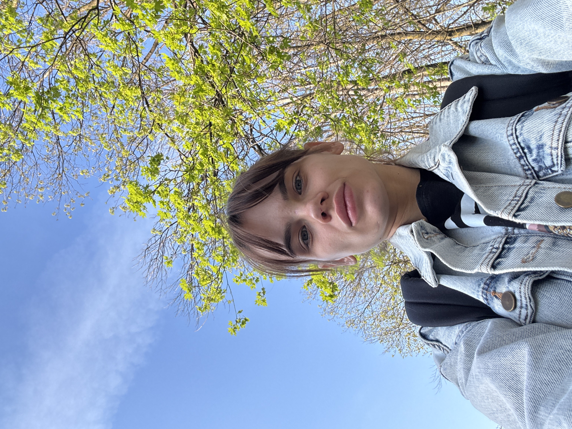 A selfie-style image captures a person with fair skin, short brown hair, and blue eyes. They are wearing a light-wash denim jacket over a black top with white markings. Behind the person is a view of tree branches with fresh green leaves set against a bright blue sky. The angle suggests the photo was taken from a slightly low perspective, looking upwards. There is no clear indication within the image to confirm it was taken in Rostov-on-Don, Russia.