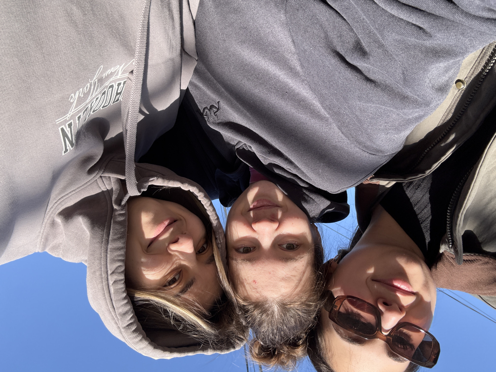 Three young women are posed closely together, looking down towards the camera. The woman on the left wears a grey hooded sweatshirt with the word "Brooklyn" printed on it. The woman in the center wears a dark grey sweatshirt. The woman on the right wears sunglasses. The background is a clear, bright blue sky. The location cannot be determined from the image, as no landmarks or other identifying features are visible.
