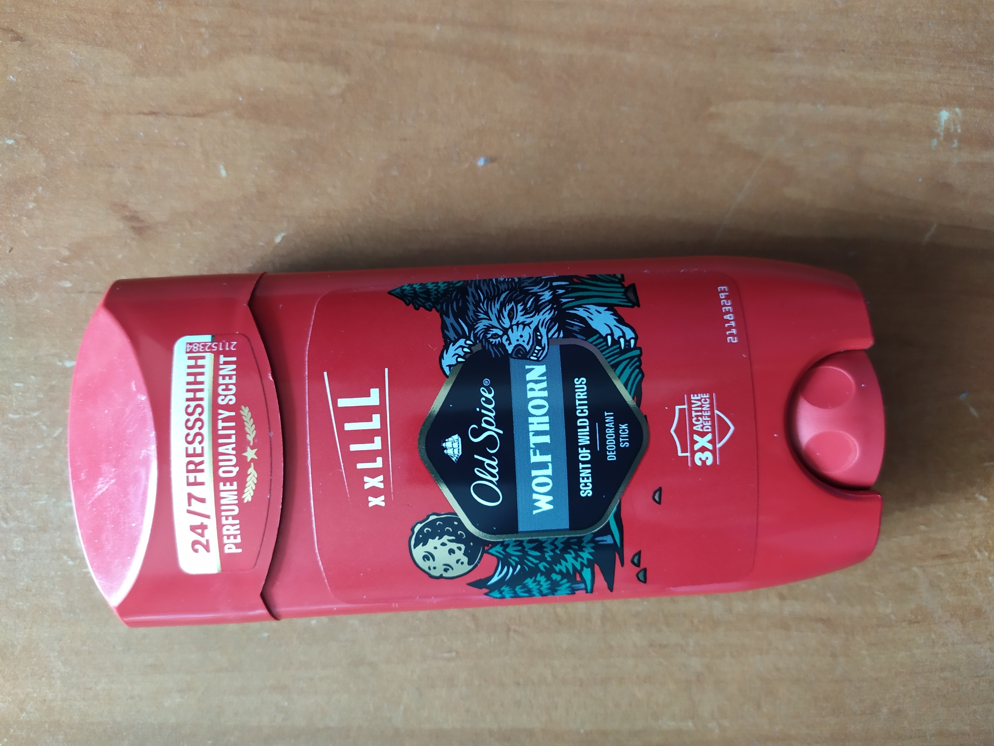 The image shows a red Old Spice Wolfthorn deodorant stick standing upright on a wooden surface. The deodorant stick features a prominent label with a wolf illustration, the Old Spice logo, and text indicating "WOLFHORN", "SCENT OF WILD CITRUS", and "DEODORANT STICK". Above this, it says "XXLLLL" and "24/7 FRESSSHHH PERFUME QUALITY SCENT". There is also a "3X ACTIVE DEFENCE" badge. The background is a textured wooden surface. The image is taken from a slightly elevated angle, looking down at the deodorant stick. There are no people or animals visible. There is no information available to indicate this image is located in the city of Turka, Ukraine.
