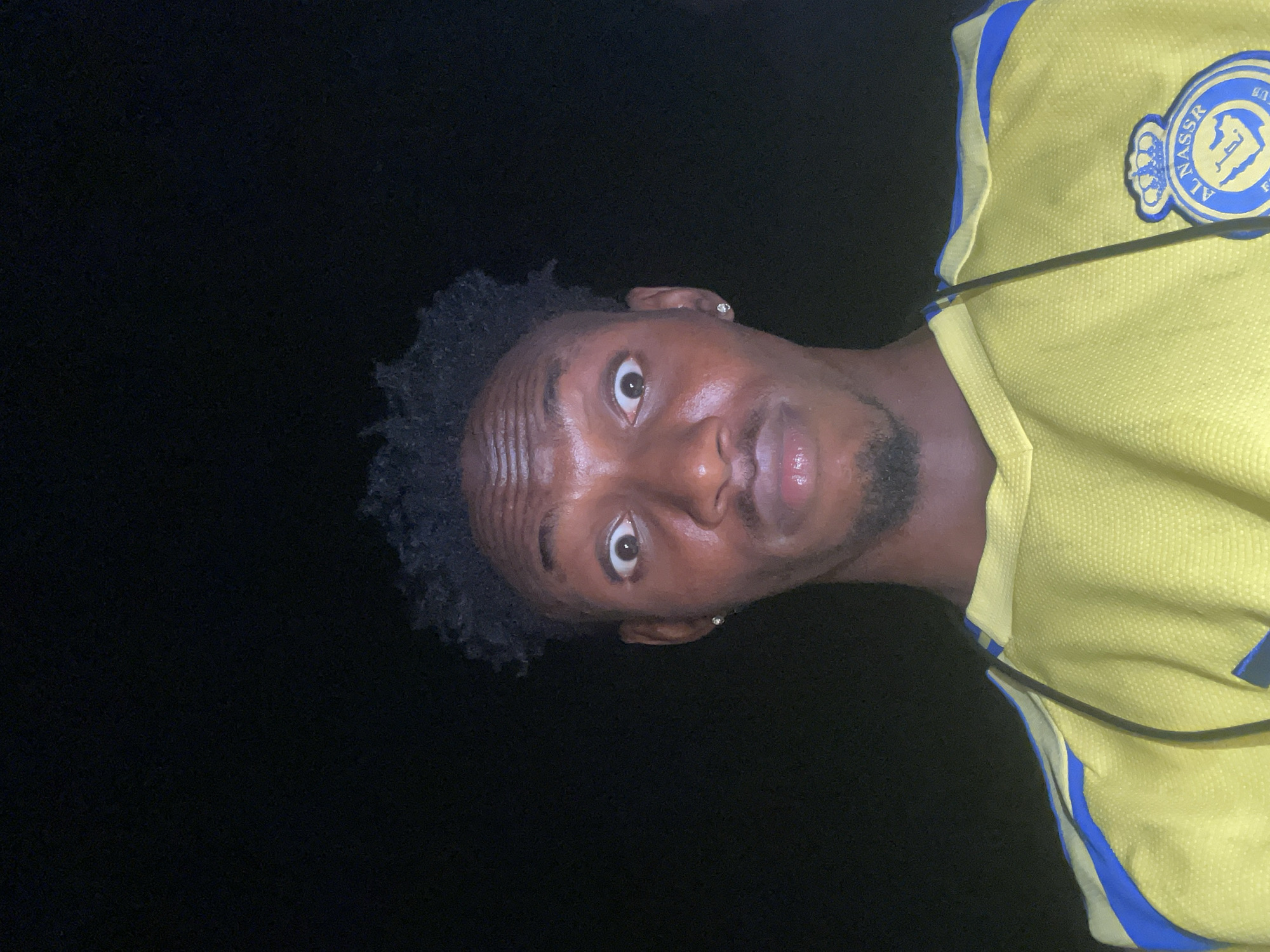 A man with dark skin and short, curly black hair is positioned in the center of the frame against a dark, blurry background. He is wearing a yellow jersey with blue accents and a logo on the upper right side of his chest. The man's face shows visible wrinkles on his forehead. His eyes are wide and looking upwards towards the camera. He has a mustache and a short beard. The lighting is dim. The image does not provide any specific details that suggest the location is Lokoja, Nigeria.