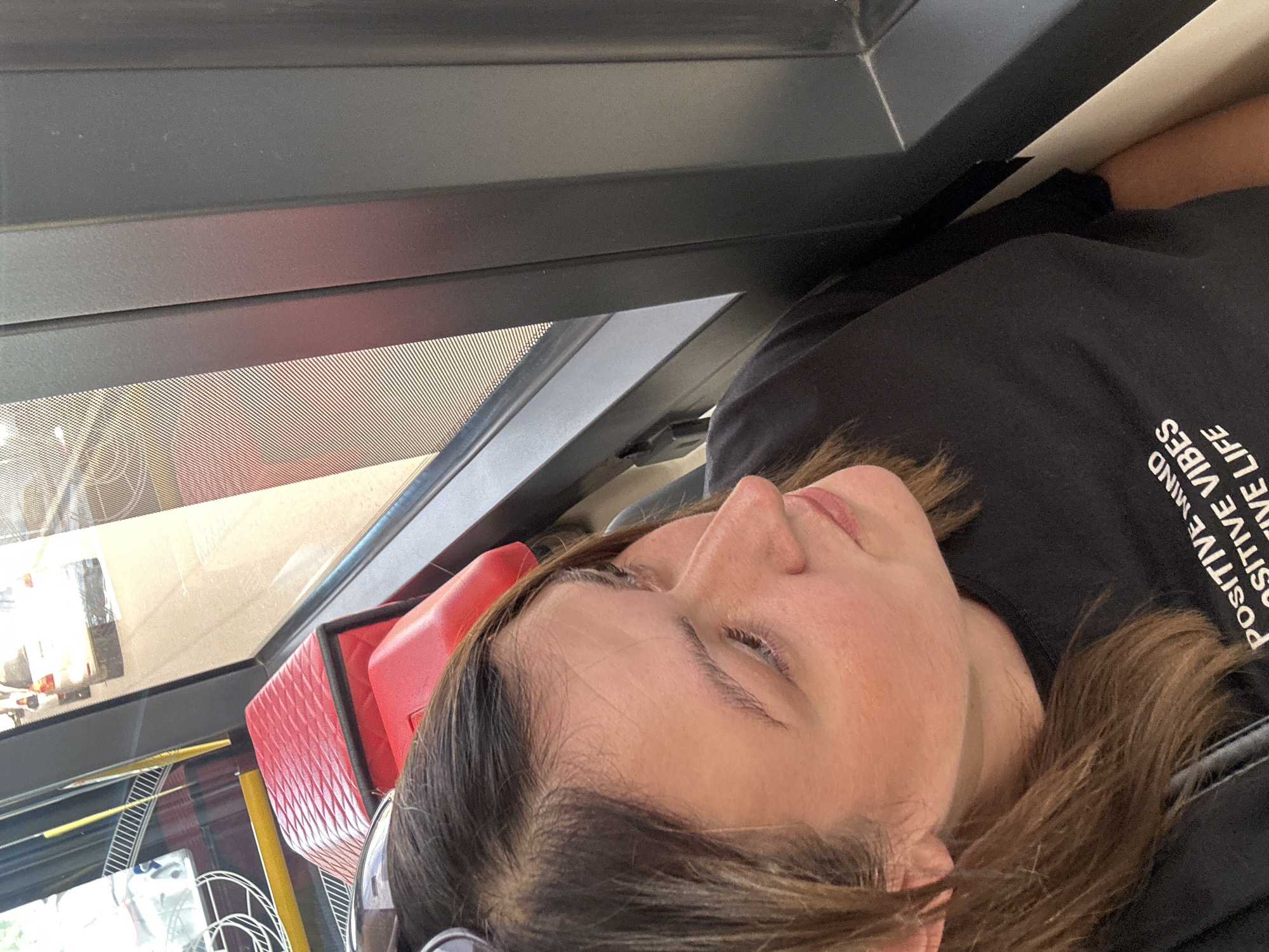 The image features a person, likely female, resting or traveling inside a vehicle, possibly a bus or train. The individual is captured in a close-up, seen from the neck up in profile, with her head tilted back and facing right. She has brown hair and a relaxed expression, with her eyes appearing partially closed or looking downwards.
She is wearing a black t-shirt with white text printed on the right shoulder and chest area. The legible text reads: "POSITIVE MIND POSITIVE VIBES POSITIVE LIFE".
The setting appears to be the interior of a conveyance, characterized by dark grey panels above a window. The window features a distinctive dotted pattern, possibly a sunshade or tint. Behind the person's head is a bright red, textured object, likely a padded seat or a headrest cover. Through the window, soft daylight illuminates the scene, and faint reflections of the exterior, possibly showing other structures or vehicles, are visible. The overall mood conveyed is one of tranquility, suggesting a moment of rest during travel. There are no specific visual cues in the image to indicate its location in Omsk, Russia.