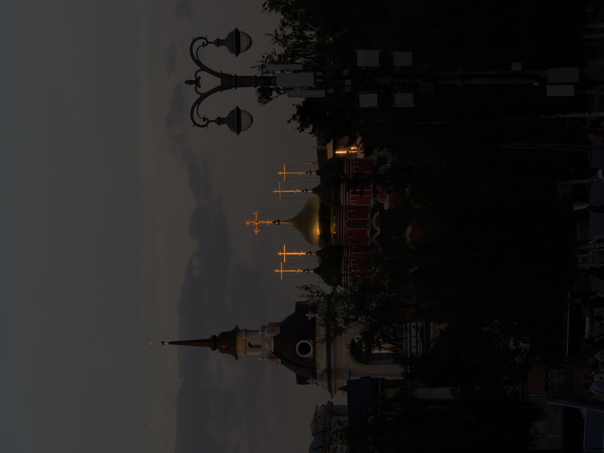 At dusk in Moscow, Russia, a vertical composition captures a street scene featuring prominent religious architecture. A lamppost stands to the left of the frame, with a complex design of curved ironwork and spherical lights.
Two churches dominate the background. The first church has a golden dome topped with a cross, with other crosses visible on adjacent buildings. The second church is characterized by a slender spire.
Trees and foliage are interspersed between the buildings. Modern communication and electrical equipment are mounted on poles or buildings. The bottom of the frame is indistinct. The sky is overcast.