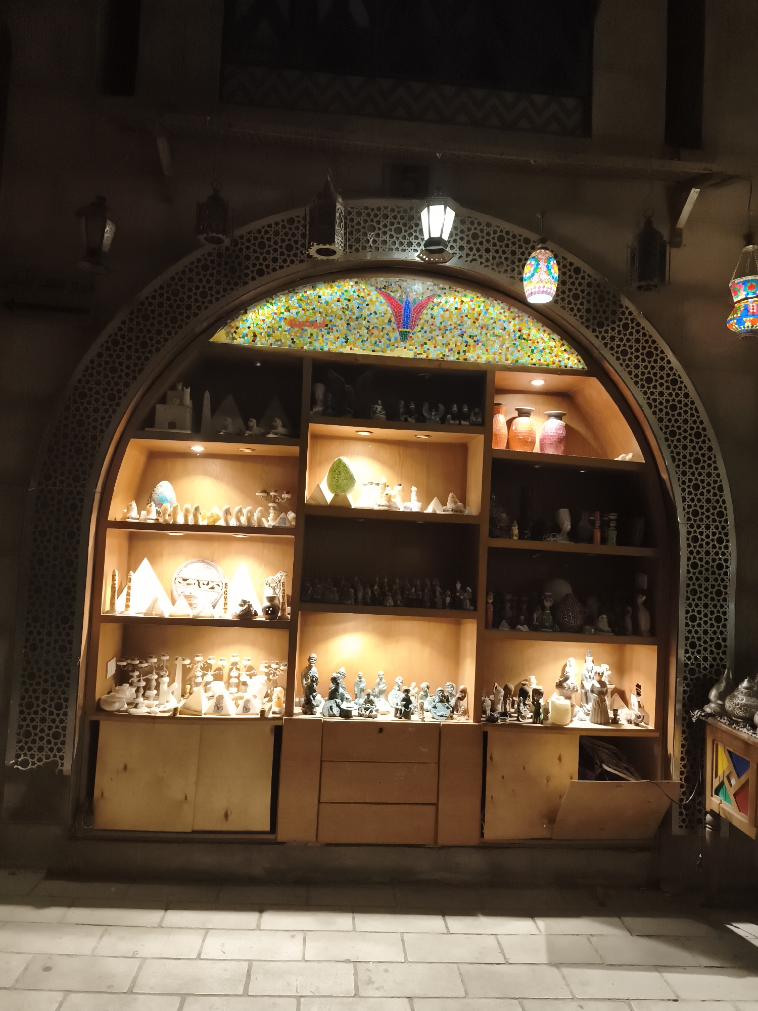 The image depicts a nighttime outdoor scene in Sharm El Sheikh, Egypt, featuring a large arched display shelf filled with various decorative items, likely souvenirs. The display unit is constructed of light-colored wood and is illuminated from within. The arched frame surrounding the shelves has an intricate geometric pattern.
The shelves are densely packed with a variety of objects, including miniature pyramids, statues resembling ancient Egyptian figures, pottery, and other decorative trinkets. Some items are lit by spotlights, highlighting their details. Above the shelves, a colorful mosaic design is visible within the arch, featuring a butterfly motif. Several decorative lanterns are hung from the ceiling or the surrounding structure, adding to the ambiance.
The foreground shows a paved walkway made of light-colored square tiles, with shadows cast by the display unit and possibly other unseen elements. The overall impression is that of a shop or market stall at night, offering Egyptian-themed merchandise. No people are visible in the image.