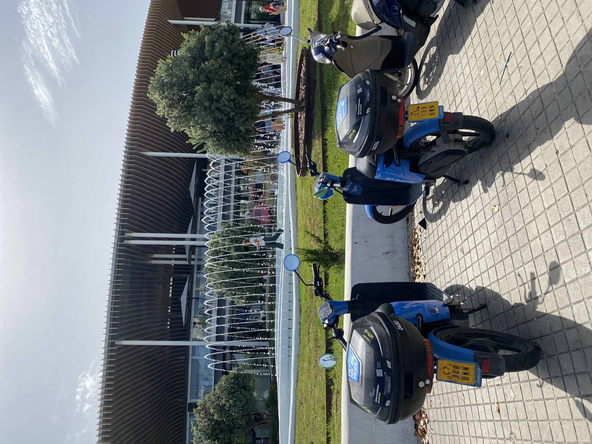 In Valencia, Spain, two blue electric scooters are parked on a paved walkway in front of a modern building with a slatted wooden facade. A fountain with multiple jets of water is located on the grass behind the scooters. Several people are visible near the fountain and inside the building. The sky is partly cloudy.