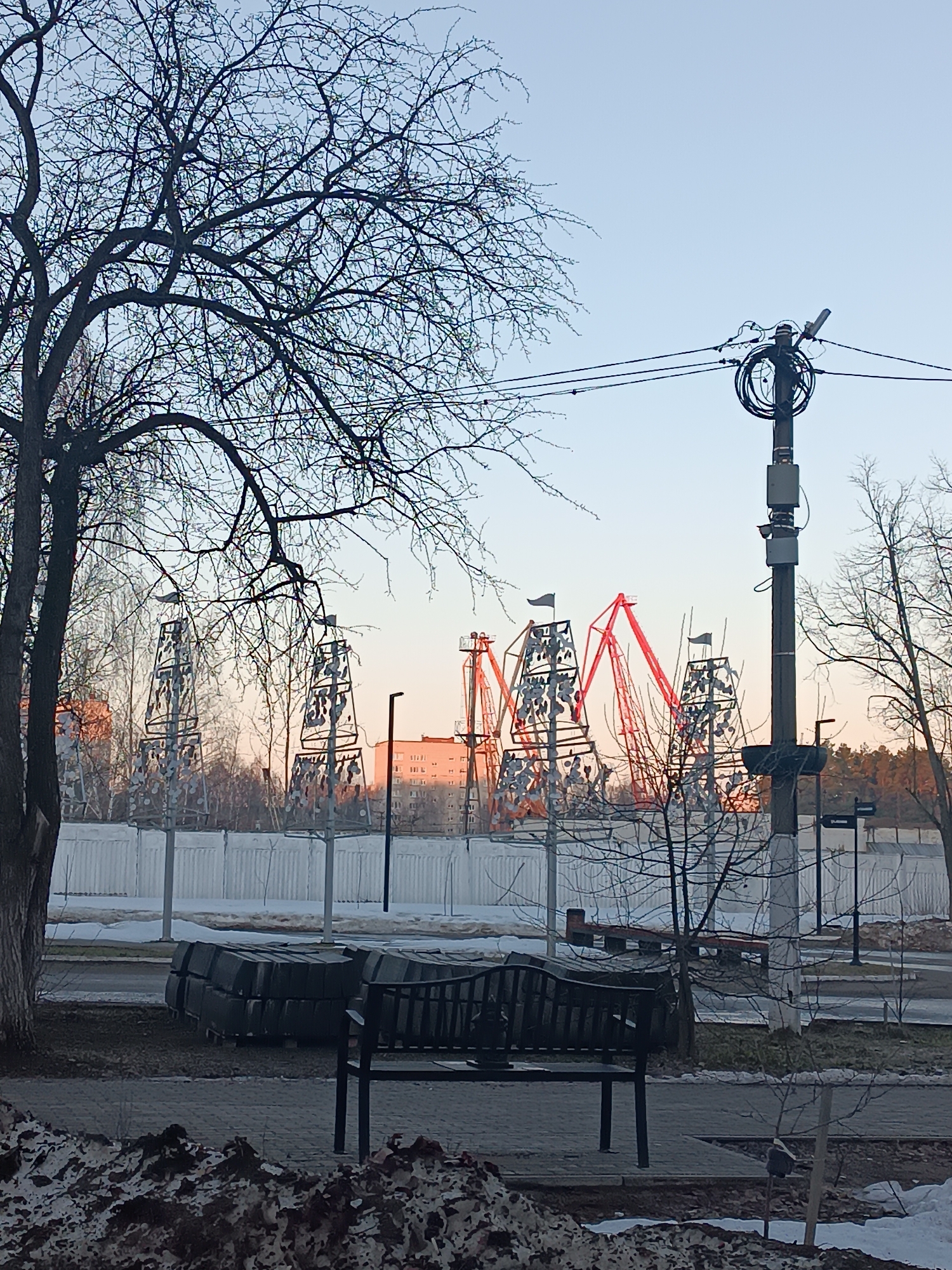 The image shows a park scene in Chaikovsky, Russia. In the foreground, there is a black metal bench with a pile of dark gray paving stones behind it. In front of the bench is a pile of snow mixed with dirt. Beyond the bench and paving stones is a paved walkway and a street with remnants of snow. A white fence runs parallel to the street. Behind the fence are several metal sculptures resembling ships, some with red accents. A multistory building is visible in the background. There are bare trees throughout the scene. A utility pole with wires and equipment attached is visible on the right side of the image. The sky is a light blue hue, suggesting either early morning or late afternoon. The overall atmosphere suggests winter or early spring.