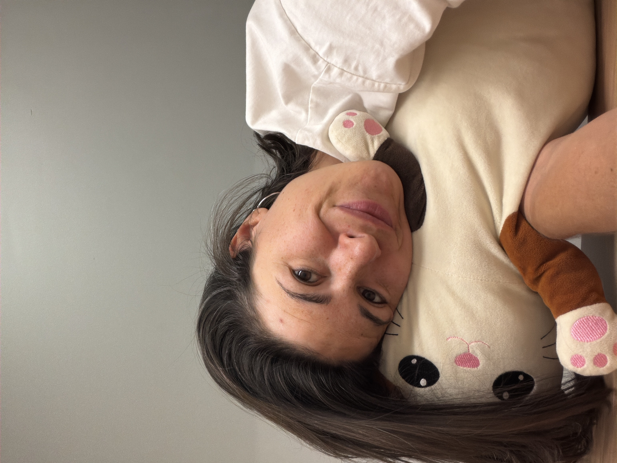 A woman with brown hair and fair skin is holding a stuffed animal. The stuffed animal is a light beige color with brown limbs and a pink nose and paws. The woman is wearing a white shirt. The background is a light gray wall. The photo is taken indoors. The location of the photo is unknown and there is no information to suggest it was taken in Budva, Montenegro.
