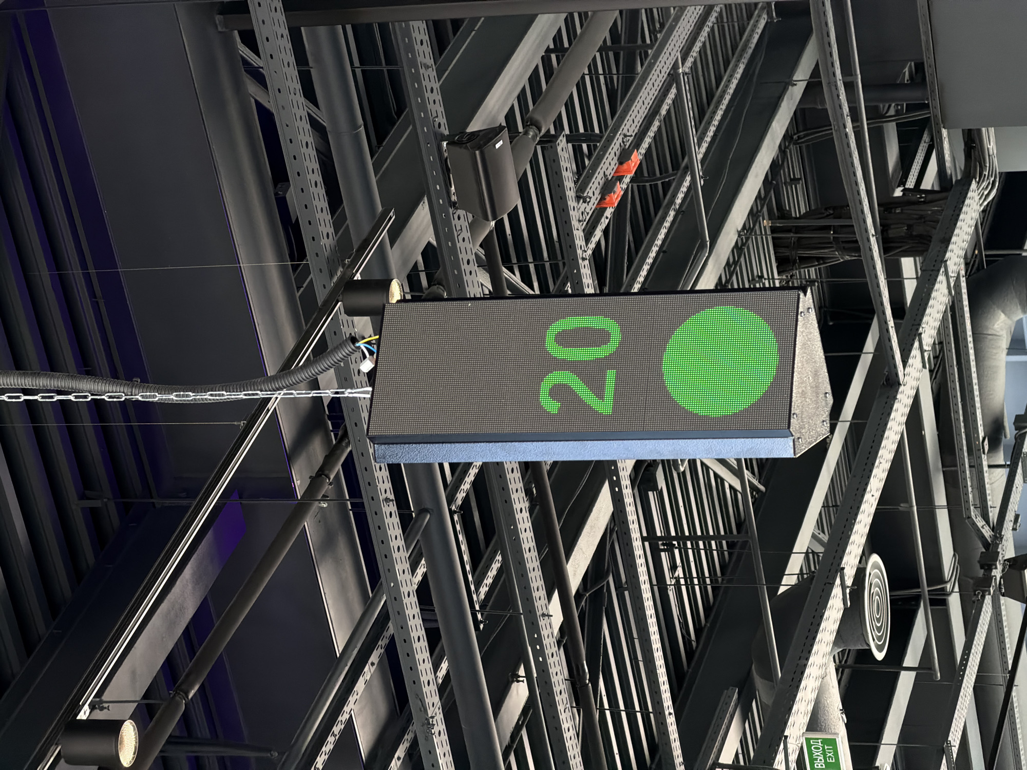 The image shows a complex network of metal beams and structures, likely part of the ceiling or support system of a large indoor space. In the foreground, a digital display shows the number "20" with a green circle next to it, hanging from the structure with a chain. Various cables and wires are visible, running along and connecting to the metal framework. A speaker is also attached to the beams. The location may be in Krasnoyarsk, Russia, although this cannot be definitively confirmed from the image alone.