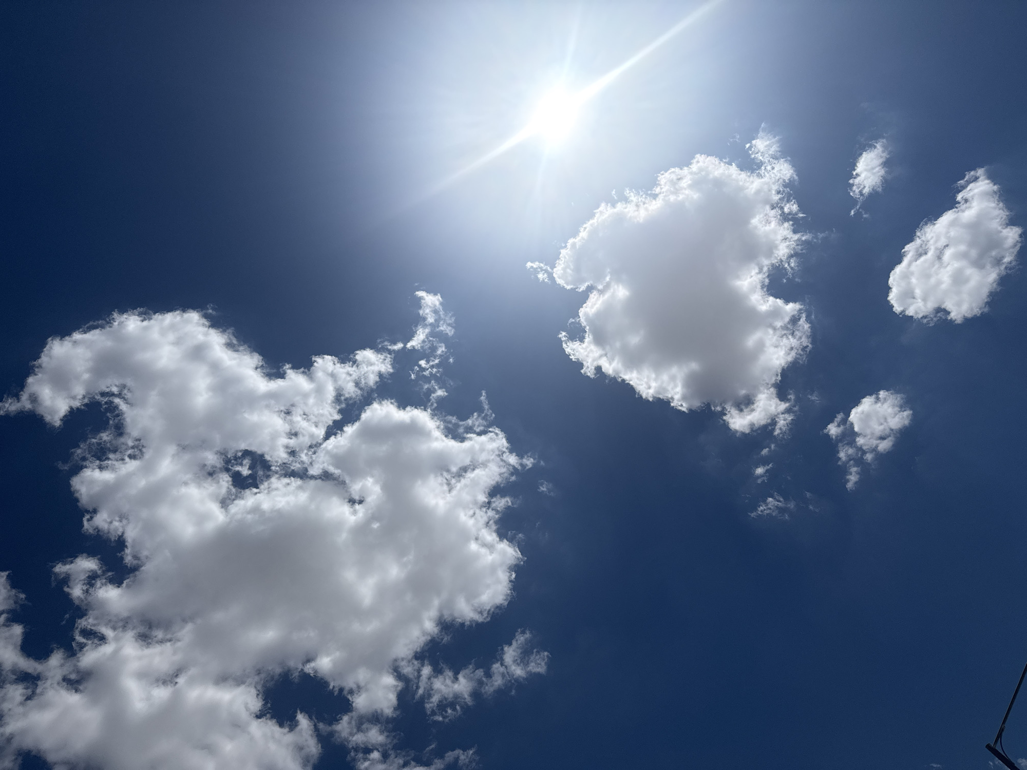 A vertical, medium shot shows a bright, sunny sky with scattered clouds. The sun, a brilliant starburst, is located in the upper right quadrant of the image, its rays fanning out across the sky. Several fluffy white clouds are dispersed throughout the deep blue expanse, some appearing dense and well-defined, while others are wispy and translucent. The lower left corner of the image shows the edge of a dark, diagonal object, possibly a pole or antenna, extending from the bottom of the frame. The overall scene evokes a clear, pleasant day with good visibility.