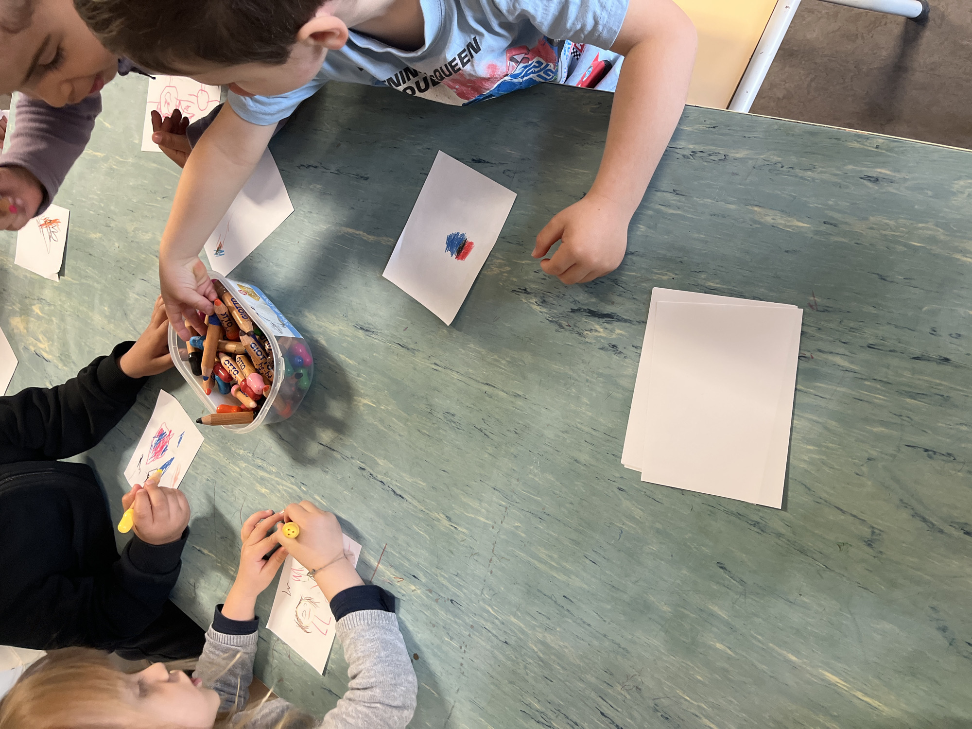 An overhead shot captures several young children engaged in an art activity at a table in an indoor setting, likely in La Fouillouse, France. The greenish-blue marbled tabletop is central to the scene, adorned with small white sheets of paper, some featuring children's drawings and scribbles.
At least four children are visible, primarily by their arms and hands. In the upper left, a child in a purple top leans over their drawing. Another child, wearing a light blue t-shirt with the partial text "...NIN QUEEN" visible, is positioned top-center/right, reaching into a clear plastic container overflowing with crayons and colored pencils, many branded "OTTO". This child has also created a small artwork with blue and red marks on a piece of paper.
To the bottom left, a child in a black long-sleeve top uses a yellow crayon to draw on their paper. Adjacent to them, a child in a grey long-sleeve top holds a colored pencil, their finger adorned with a yellow smiley face ring or holder, as they work on their own creation. A stack of fresh, blank white paper is neatly placed on the right side of the table, ready for more drawings. The children appear focused and absorbed in their creative activity. A white chair or table leg is partially visible in the upper right background.