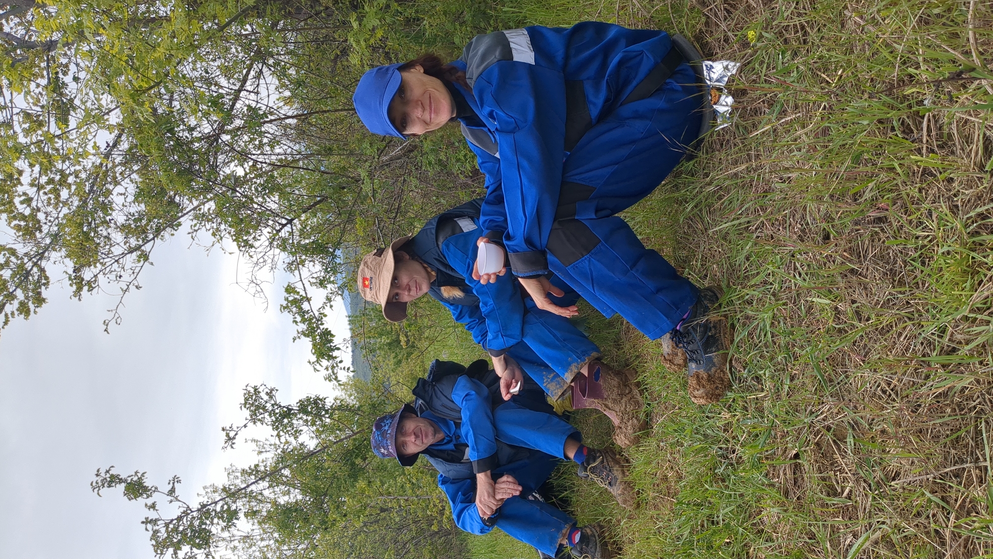 Three people are in a grassy area with trees and a cloudy sky. All three individuals are wearing blue jumpsuits. The person closest to the camera is sitting with their knees bent, wearing a patterned hat. The person in the middle is holding a small white object, and their footwear is covered in mud. The person furthest from the camera is wearing a blue cap. The location is in a natural setting, not a city. It is not possible to confirm that this photo was taken in Pushkino, Ukraine.