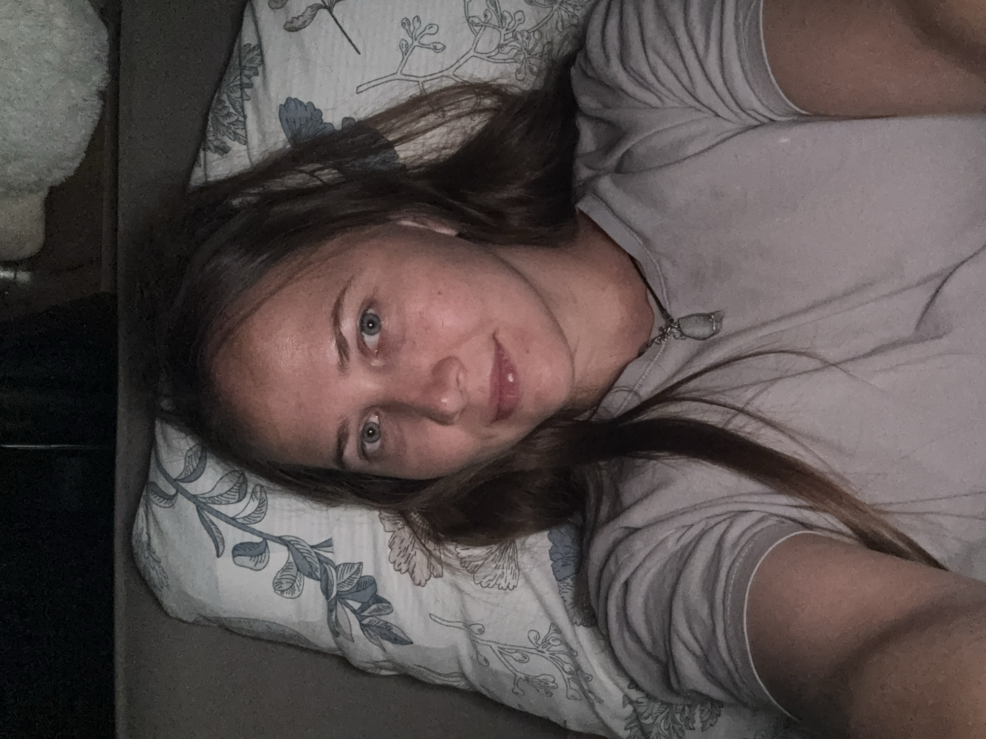 A woman with long, brown hair is lying in bed, looking at the camera. She is wearing a light gray t-shirt and a necklace with a pendant. The pillow she is resting her head on has a blue and white floral pattern. The background is dark, suggesting the room is dimly lit. The image appears to be a selfie taken indoors. There is no specific information in the image that would allow for identification of a location in Izhevsk, Russia.