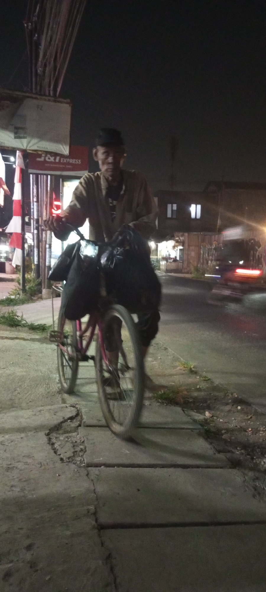 A man wearing a dark cap and a light-colored long-sleeved shirt rides a bicycle at night in Setu, Indonesia. He is carrying two large black bags, one on each side of the handlebars, and appears to be on a sidewalk. The bicycle has a pink frame and is moving. The background shows a dimly lit street with some buildings and a vehicle in motion. The sidewalk is cracked and uneven.