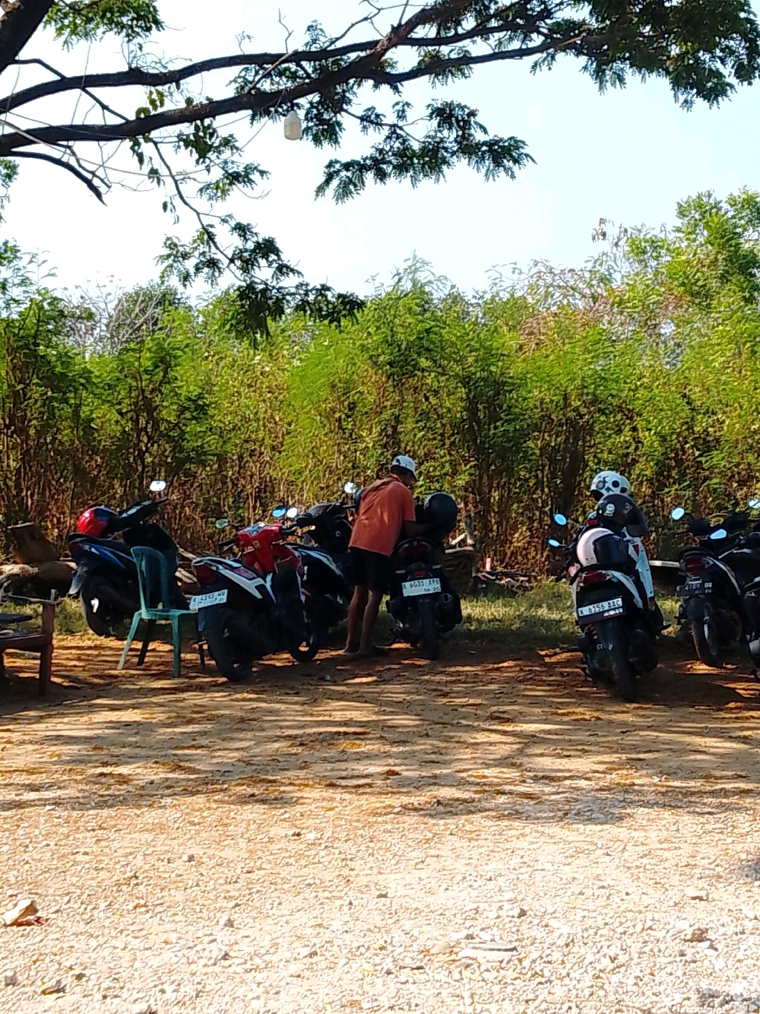 The image depicts a dirt parking area with several motorcycles lined up under a canopy of trees. A person wearing an orange shirt and shorts is standing next to a motorcycle. Another person wearing a helmet is seated on a motorcycle in the background. The environment appears to be outdoors with dense vegetation in the background, suggesting a rural or semi-rural setting. The scene is lit by natural sunlight, creating shadows on the ground. The image was captured in Cikal, Indonesia.