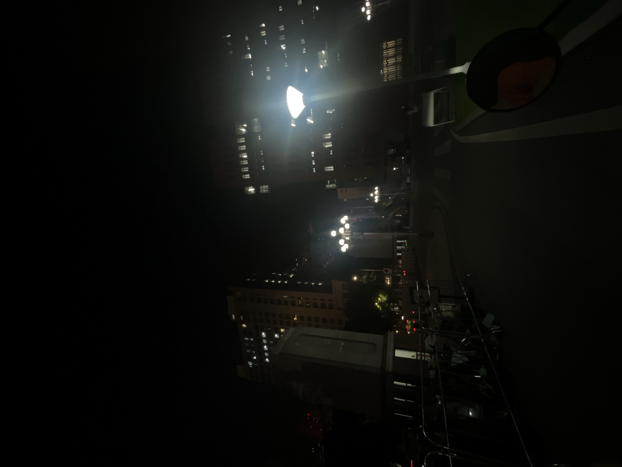 The image depicts an urban environment at night in Yuzhong District, China. The scene is dominated by artificial lighting from streetlights and illuminated windows in multi-story buildings. In the lower right foreground, a circular rearview mirror is visible, partially reflecting an orange object. A paved pathway or road occupies the center and right foreground, marked by a curved white line and a straight white line, with a narrow strip of green landscaping alongside. To the left of the pathway, a section of metal barriers, possibly bicycle racks, encloses several dark shapes resembling parked bicycles or scooters.
In the midground, towards the left, a tall, rectangular stone monument or tablet with dark text stands prominently. Behind it and further left, a large, light-colored building rises, displaying numerous illuminated rectangular windows across multiple floors. To the right of the monument, a cluster of streetlights with multiple spherical globes emits a warm yellow light. Further right, a modern streetlight with a single, upward-facing conical fixture shines brightly with white light, lens flare emanating from it. Behind this streetlight, another large, multi-story building fills the right side of the background, also featuring numerous illuminated rectangular windows. A faint human figure is discernible on the pathway near the base of this building. The sky is dark, and the overall environment is dimly lit except for the concentrated areas around the light sources.