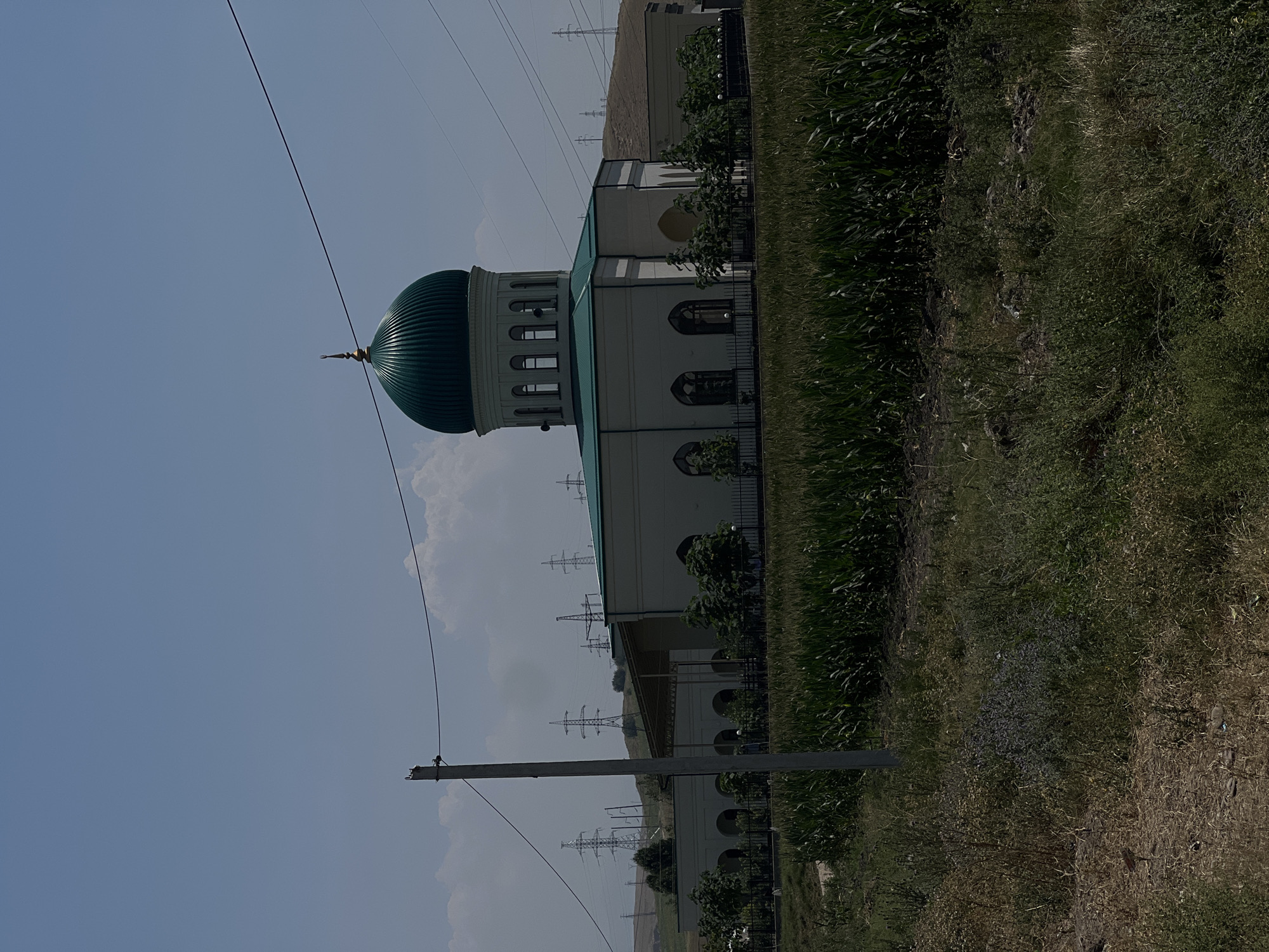 The image shows a mosque with a green dome and a green roof, situated in Karamanas, Uzbekistan. The mosque is a light-colored building with arched windows and is surrounded by a black fence. In the foreground, there is a field of tall green crops, likely corn. To the left of the mosque, a wooden utility pole stands with wires extending across the frame. In the background, several electricity pylons are visible on a hilly landscape under a hazy blue sky with scattered clouds. The overall scene depicts a rural or semi-rural setting. There are no visible people or specific actions taking place.